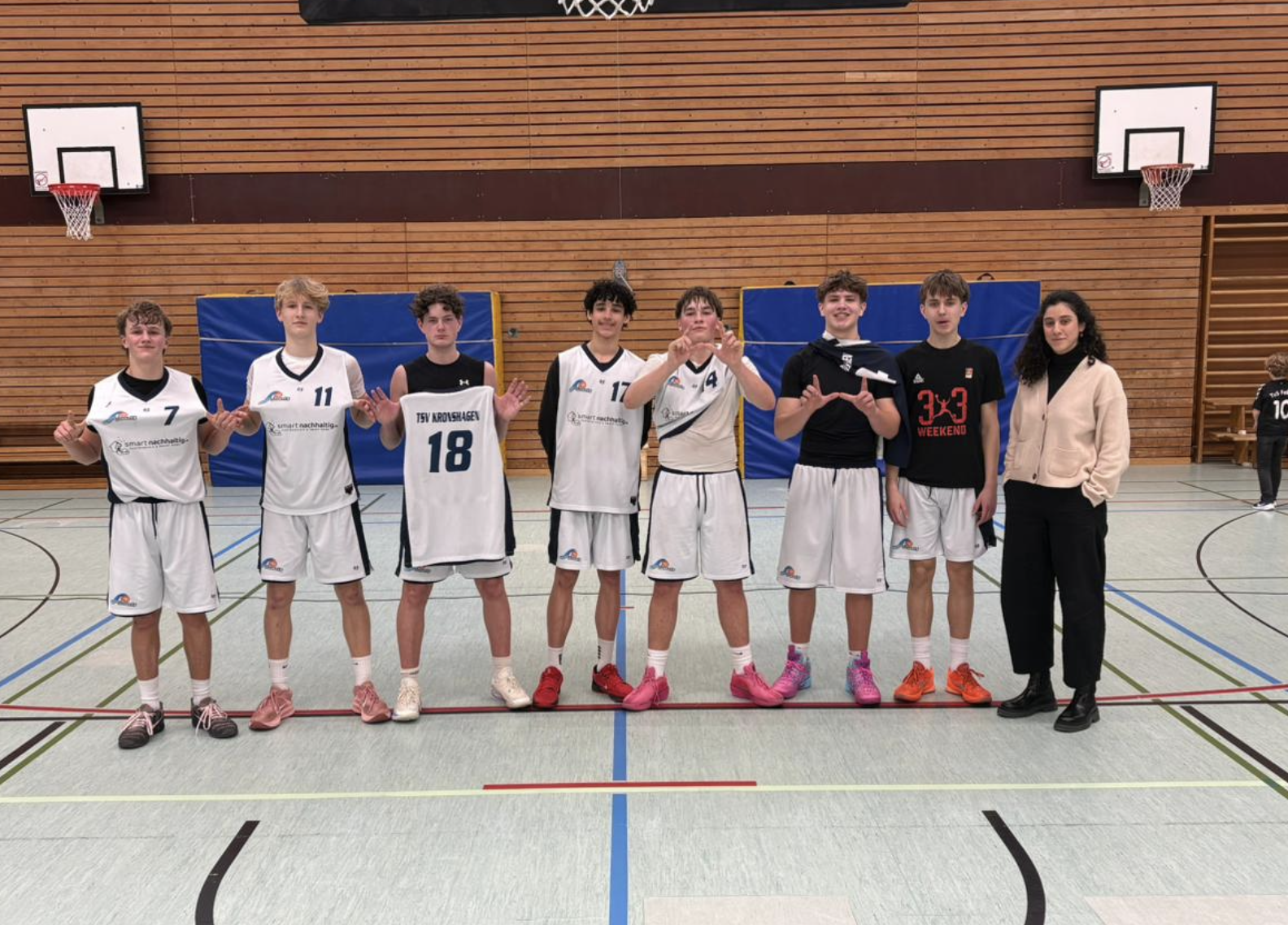 Jugendbasketballteam in Turnhalle pose mit Trainerin, alle in weißen Trikots, einige mit schwarzen oder roten Schuhen, hinter ihnen eine Holzverkleidung an der Wand.