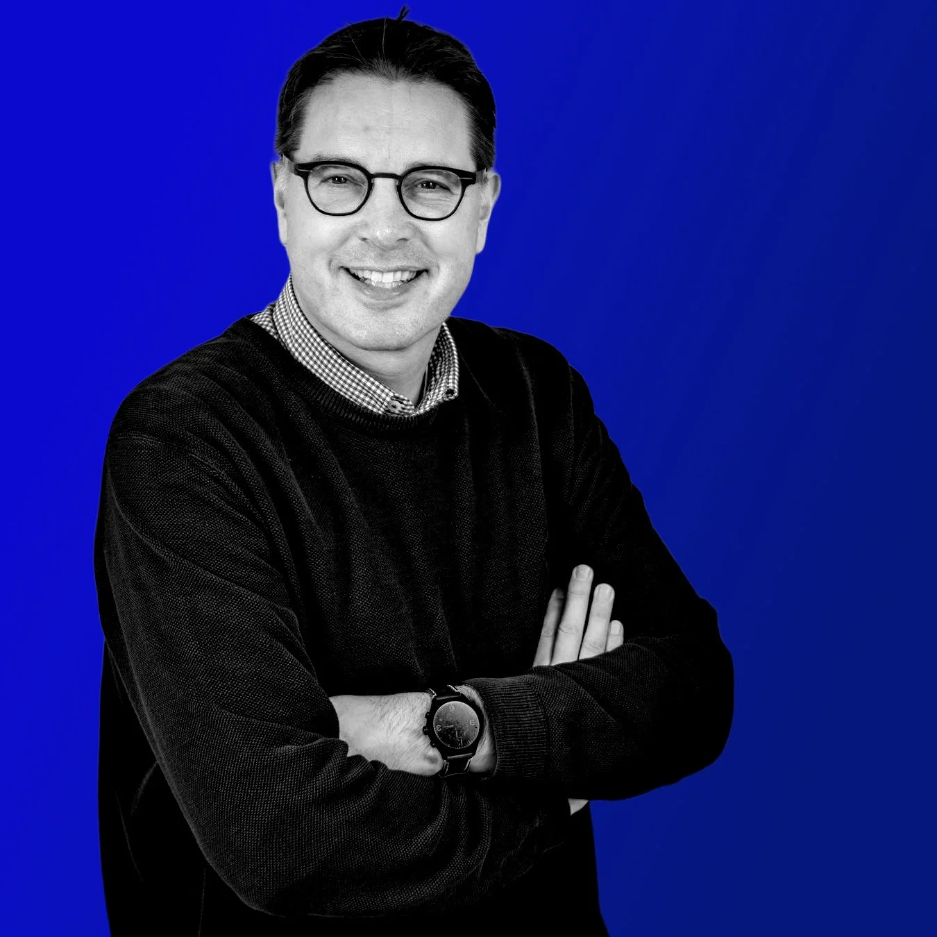 Ein Mann trägt eine Brille, trägt einen schwarzen Pullover und hebt die Arme verschränkt vor einem blauen Hintergrund. Er lächelt.