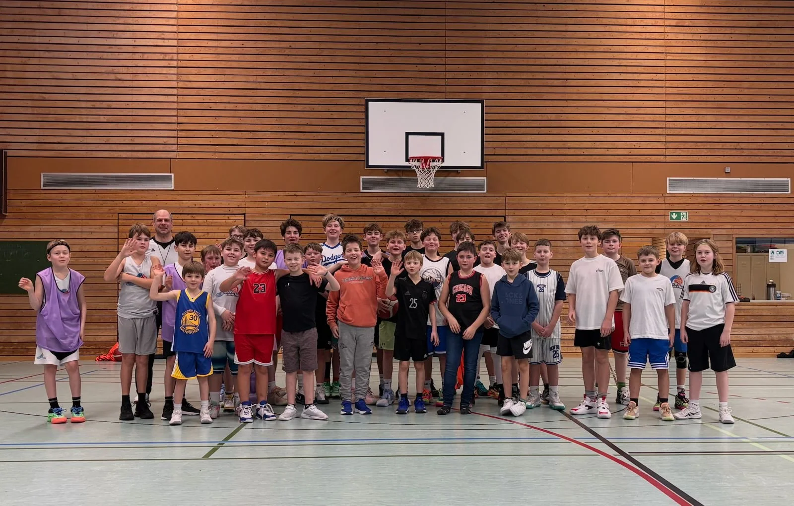 Gruppe von Kindern und einem Erwachsenen in Turnhallen, alle lächelnd, mit Basketballkörben im Hintergrund.