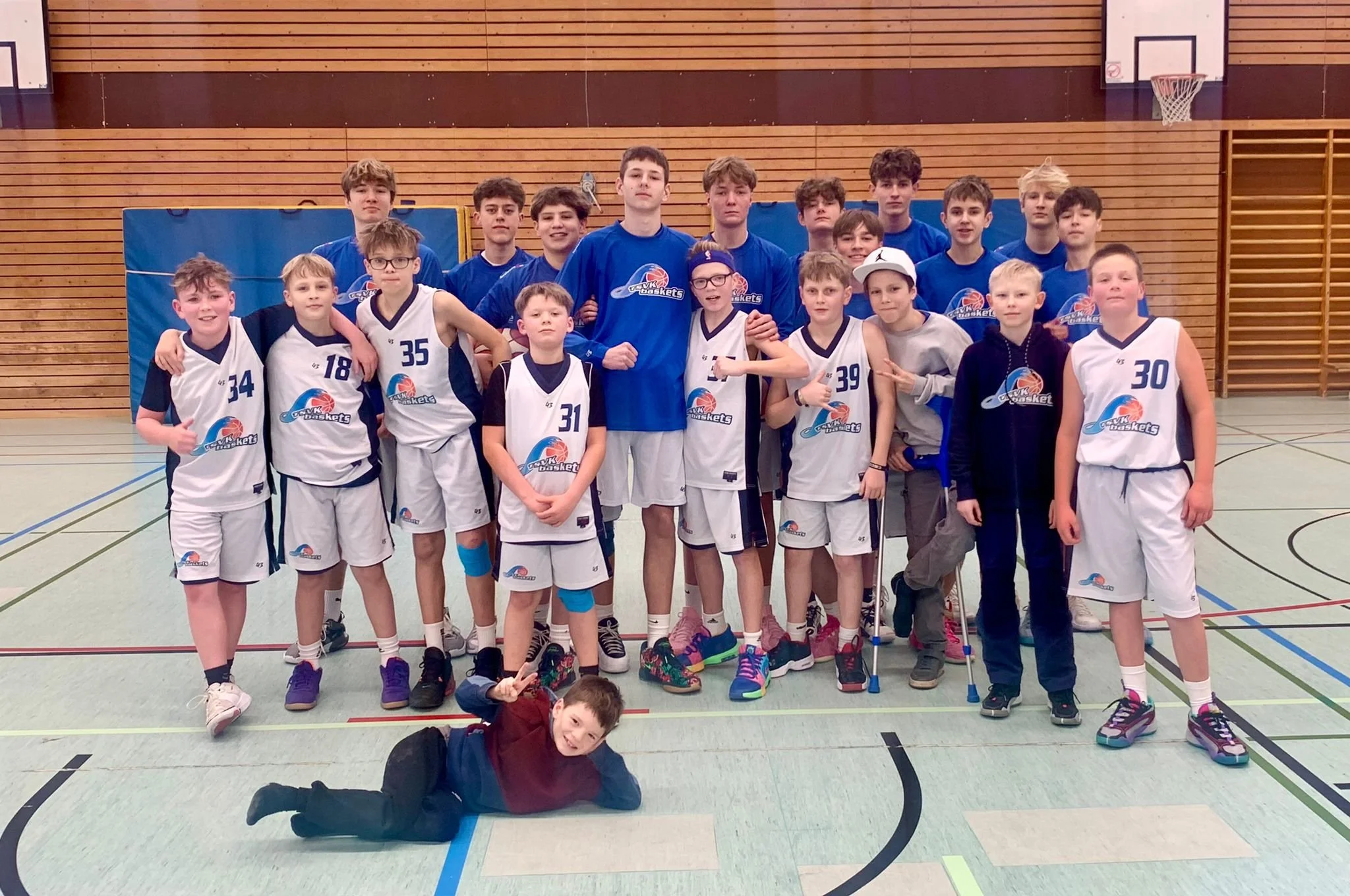 Eine Gruppe von jungen Basketballspielern und Jugendlichen steht in einer Turnhalle, einige tragen Trikots mit der Aufschrift "OVK Basket". Es sind 17 Personen zu sehen, die meisten tragen blaue Trikots, einige in weißen Trikots und eine Person trägt