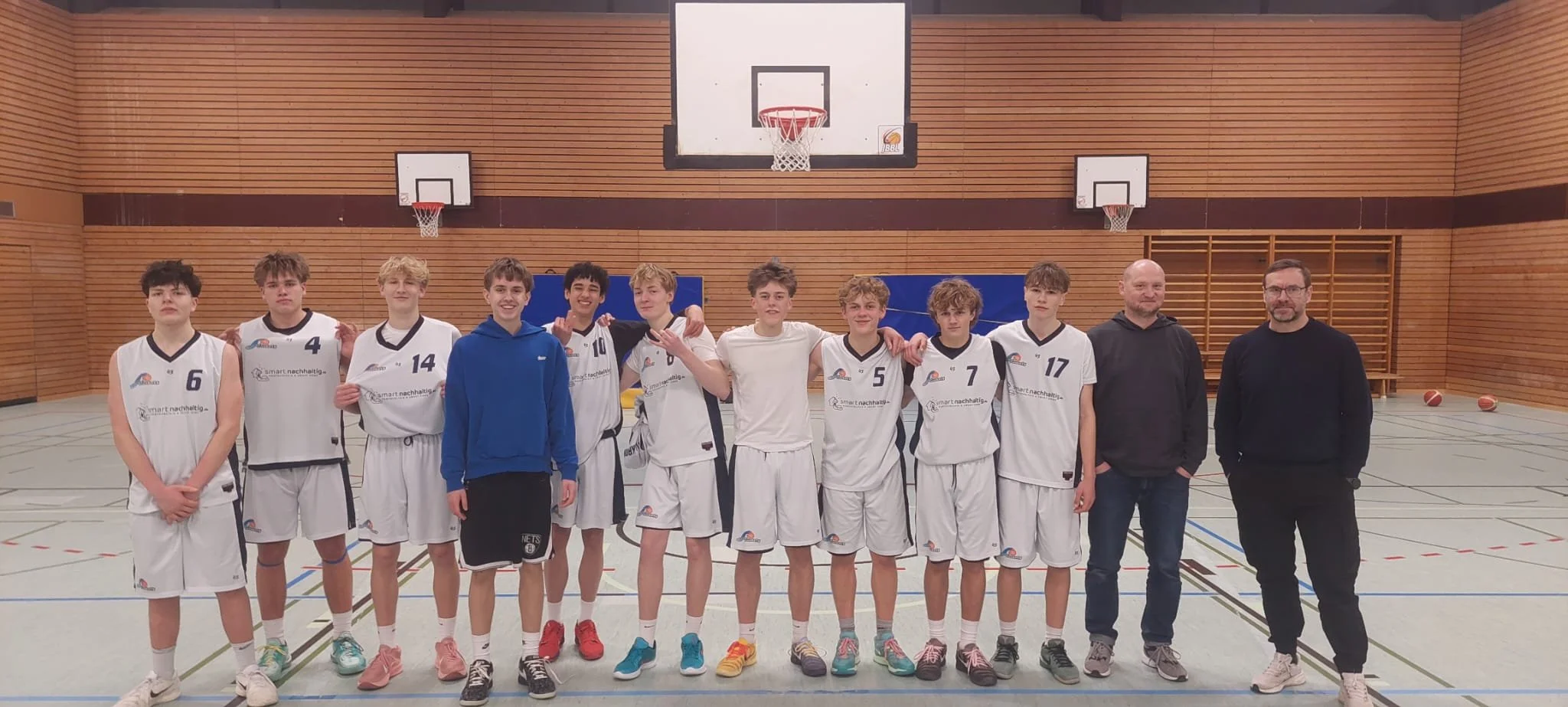 Jugendbasketballteam mit Trainern steht im Basketballhalle, alle in weißen Trikots, einige mit Nummern, Gruppe lächelt und macht Fotos, Basketball im Hintergrund.