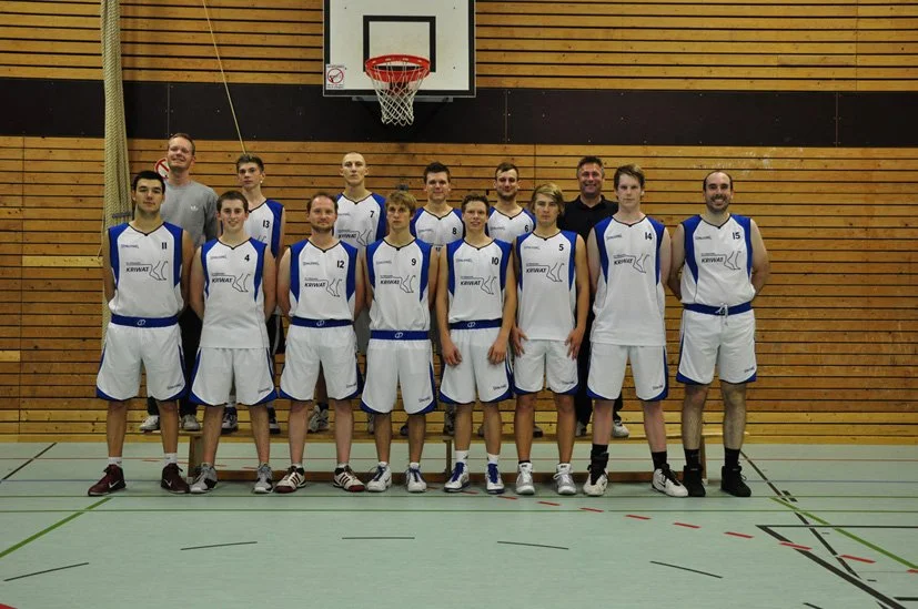 Basketballmannschaft in weißen Trikots und kurzen Hosen posiert in einer Turnhalle vor einer Holzwand, mit einem Basketballkorb im Hintergrund.