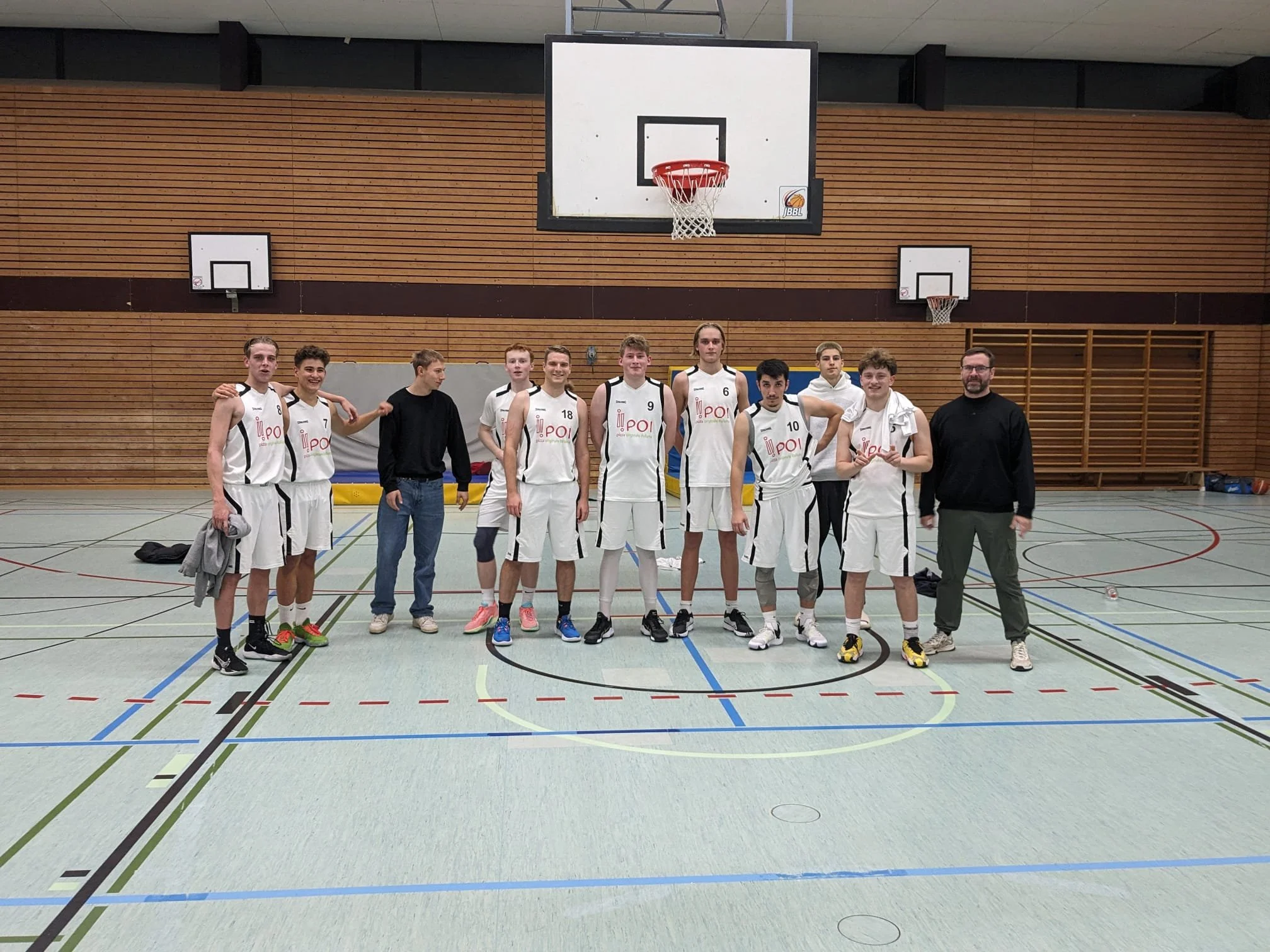 Eine Gruppe von zehn männlichen Basketballspielern und einem Mann in Zivilkleidung steht auf einem Basketballfeld in einer Turnhalle, alle lächelnd in die Kamera schauend.