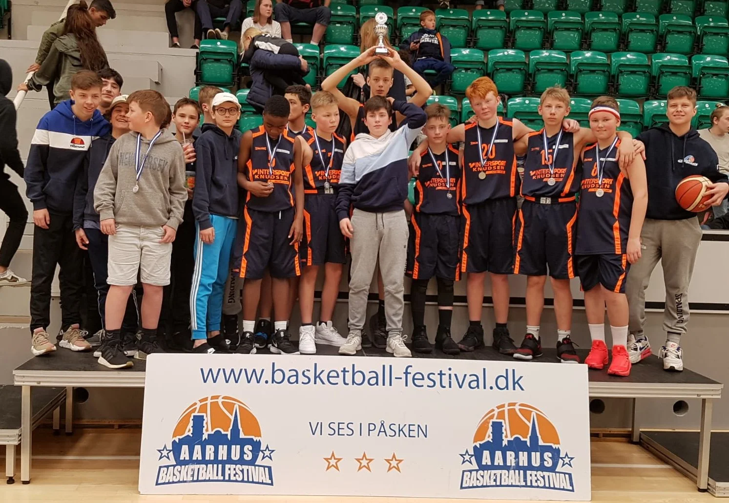 Eine Jugendbasketballmannschaft feiert ihren Sieg auf einem Podest, mit einem Trainer, die Medaillen um den Hals, und eine Trophäe in der Hand, vor einem Banner des Aarhus Basketball Festivals.