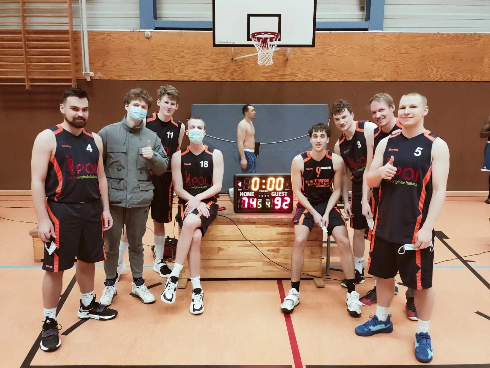 Eine Gruppe von sechs jungen Männern in Basketball-Uniformen mit Nummern und Masken, die in einer Turnhalle vor einer Basketballtafel und einem Spielstand-Display stehen. Sie scheinen nach einem Spiel zu posieren.
