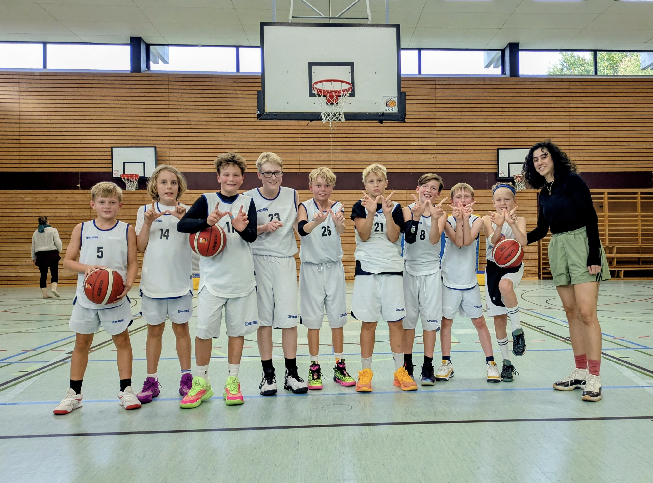 Gruppe von jungen Basketballspielern mit einer weiblichen Trainerin in einer Turnhalle. Die Kinder tragen Basketballuniformen und Schuhe, einige halten Basketballbälle. Sie posieren für das Foto, während im Hintergrund eine Holzwand und Basketballkör