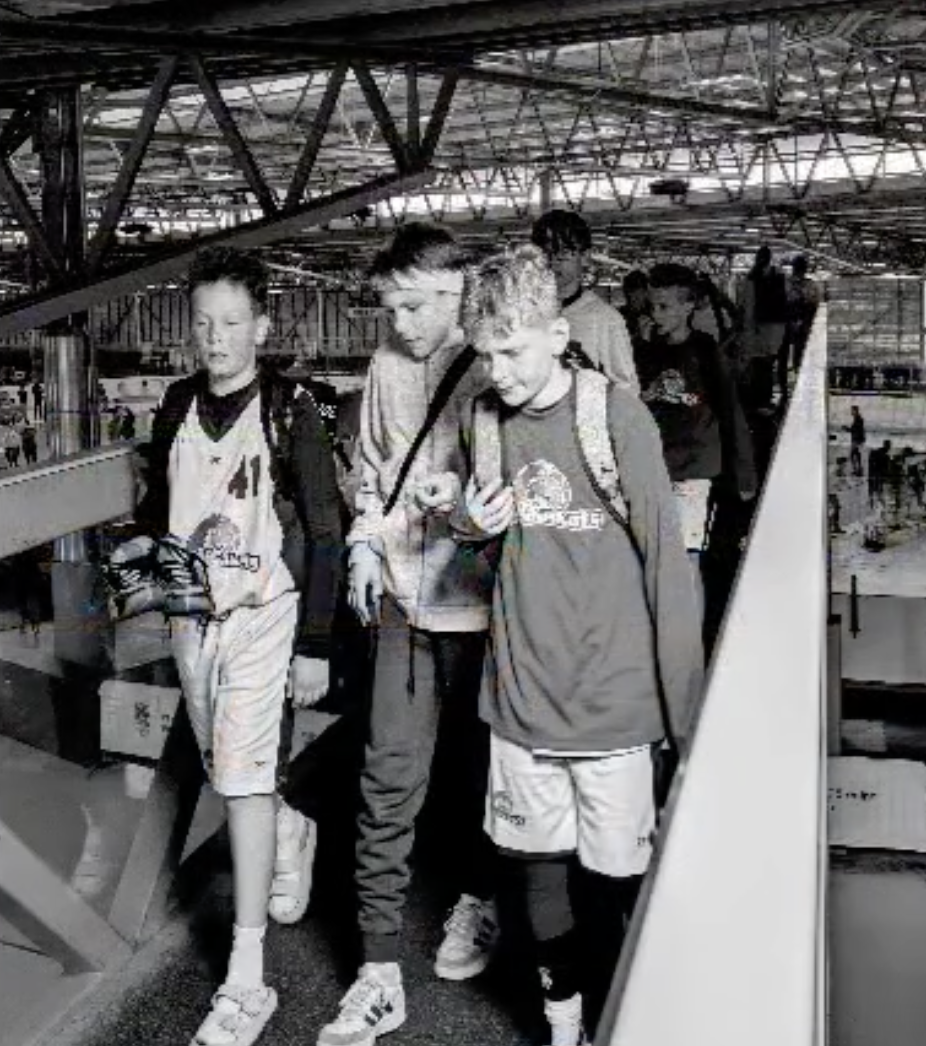 Jugendliche auf einer Brücke im Flughafen, drei Jungen sind im Vordergrund, einer schaut auf sein Handy, alle tragen Rucksäcke.