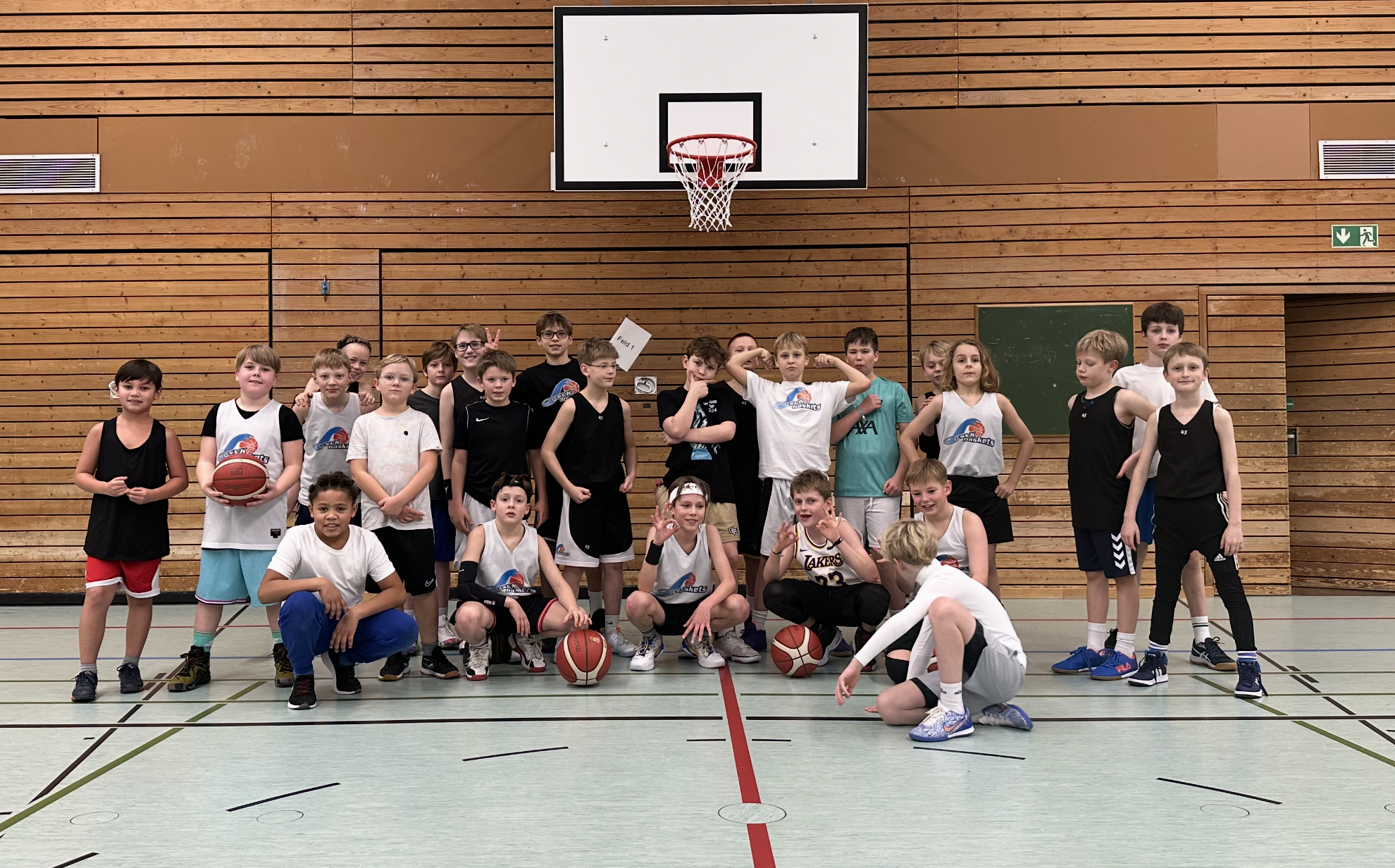 Eine Gruppe von Jungen und Mädchen in Basketballtrikots und Sportkleidung, die in einer Turnhalle mit Holzverkleidung stehen, einige sind sitzend auf dem Boden, andere stehen und posieren.