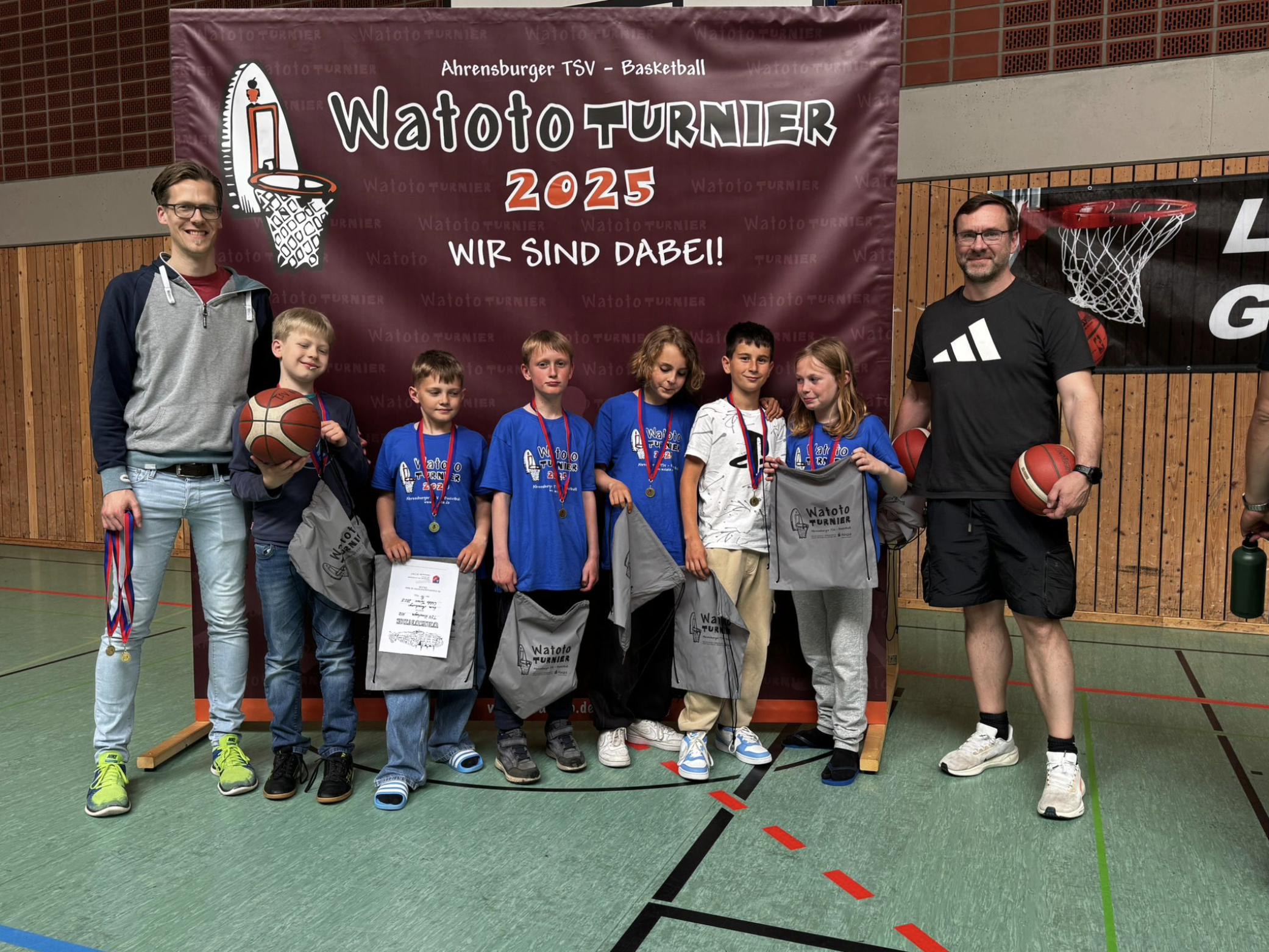 Gruppenfoto von Kindern, Trainern und Betreuern bei einem Basketballturnier, vor einem roten Banner mit der Aufschrift »Waoto Turnier 2025«. Die Kinder tragen Medaillen und halten Geschenke, während die Betreuer lächeln.