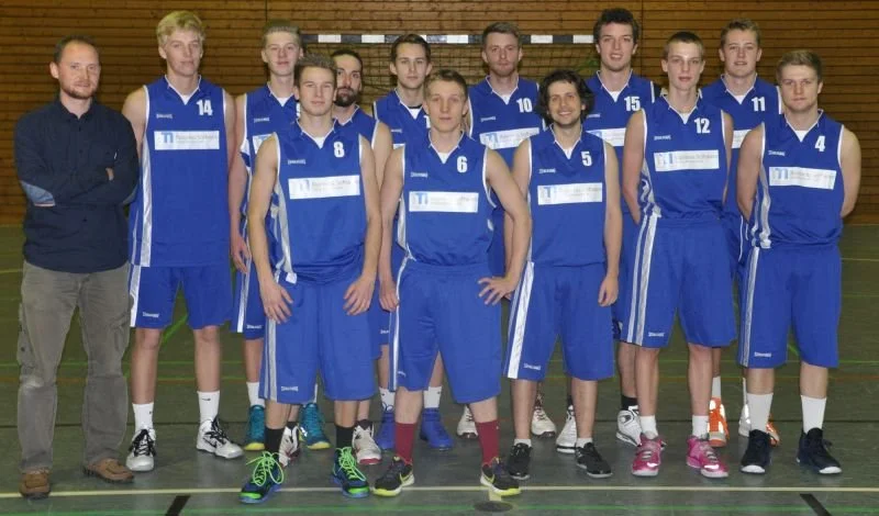 Eine Gruppe junger Basketballspieler in blauen Trikots und ein Mann in dunklem Hemd stehen in einer Halle, wahrscheinlich vor einem Basketballspiel oder -training.