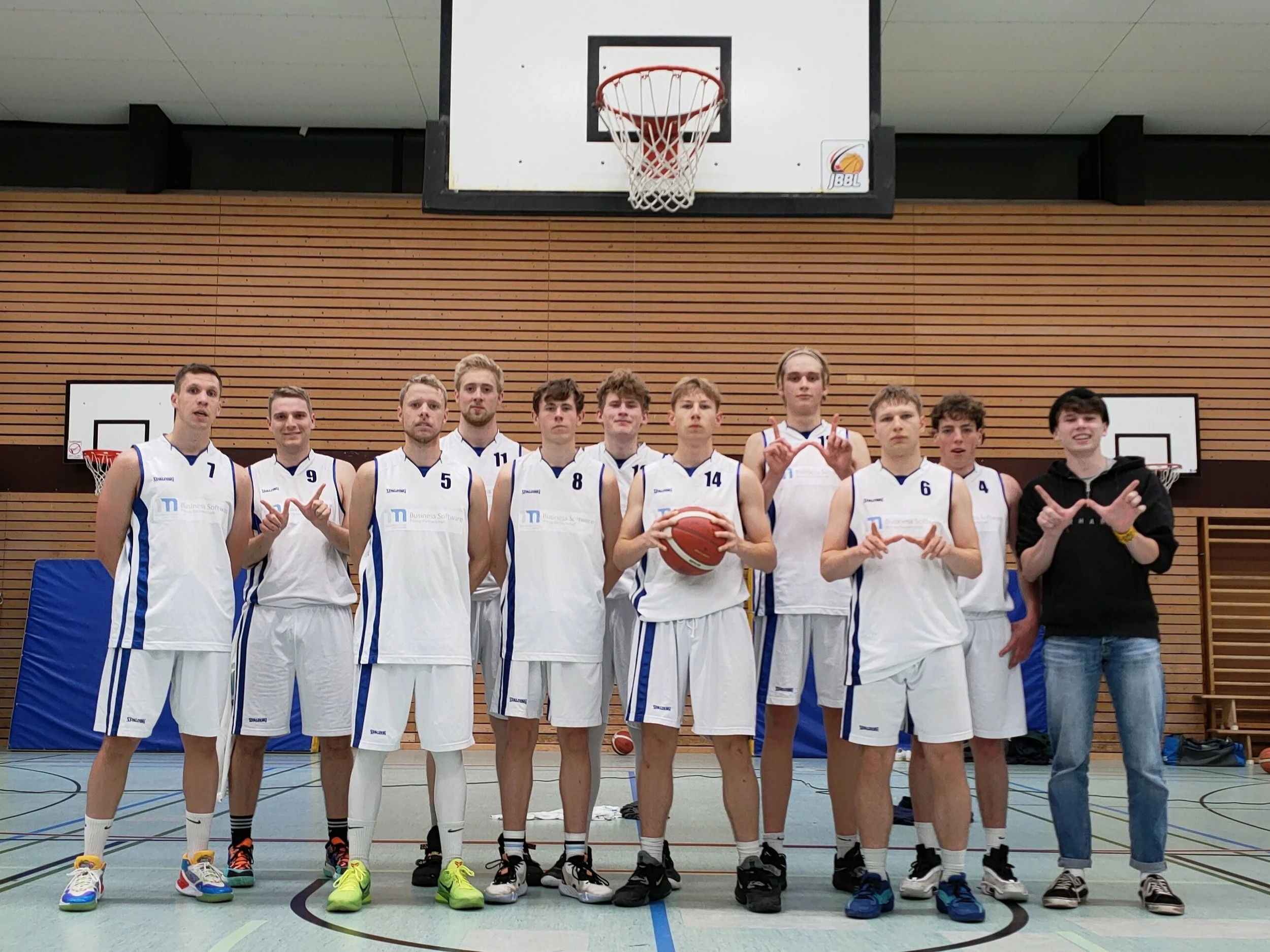 Ein Basketballteam in weißen Trikots steht in einer Halle, alle schauen in die Kamera, mit einem Basketball in der Hand, im Hintergrund Basketballkörbe an der Wand.