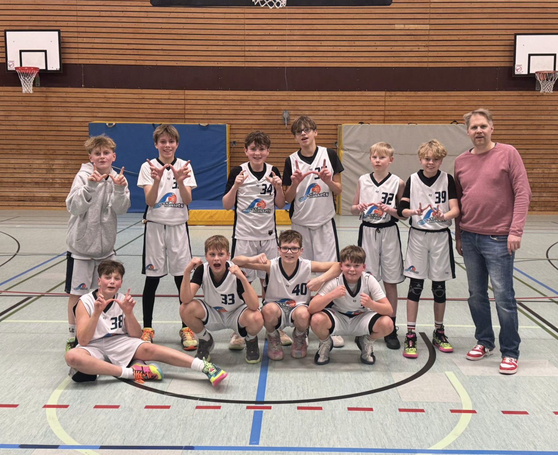 Eine Gruppe junger Basketballspieler und ein erwachsener Trainer posieren in einer Sporthalle für ein Foto. Die Kinder tragen weiße Trikots mit schwarzen Ärmeln und sitzen oder stehen vor einem Holz-Wands des Turnhalls, auf dem Basketballkörbe zu seh