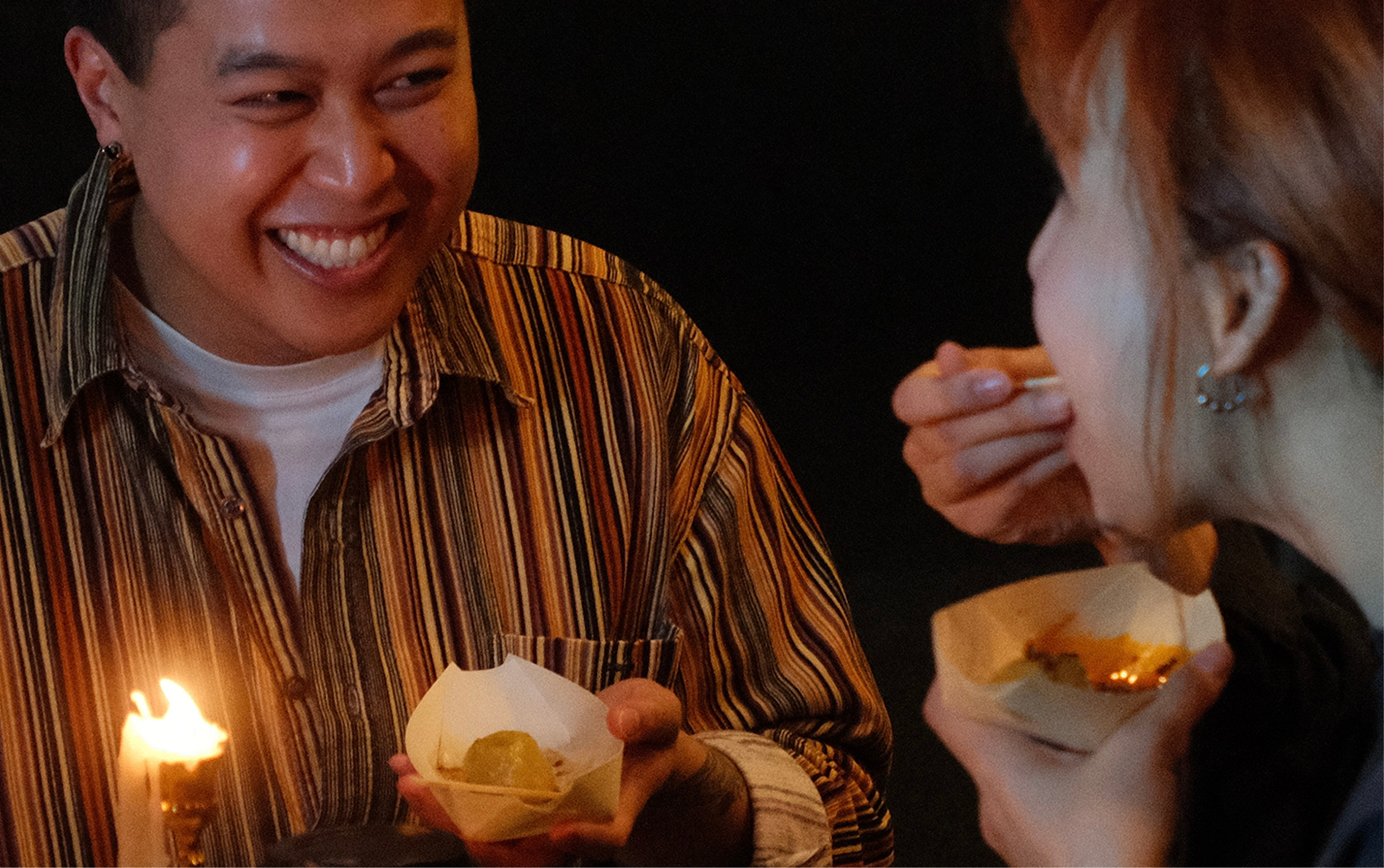 Two people smiling while eating and tasting Fuzhounese potato balls from small paper trays in a warm, cozy setting.