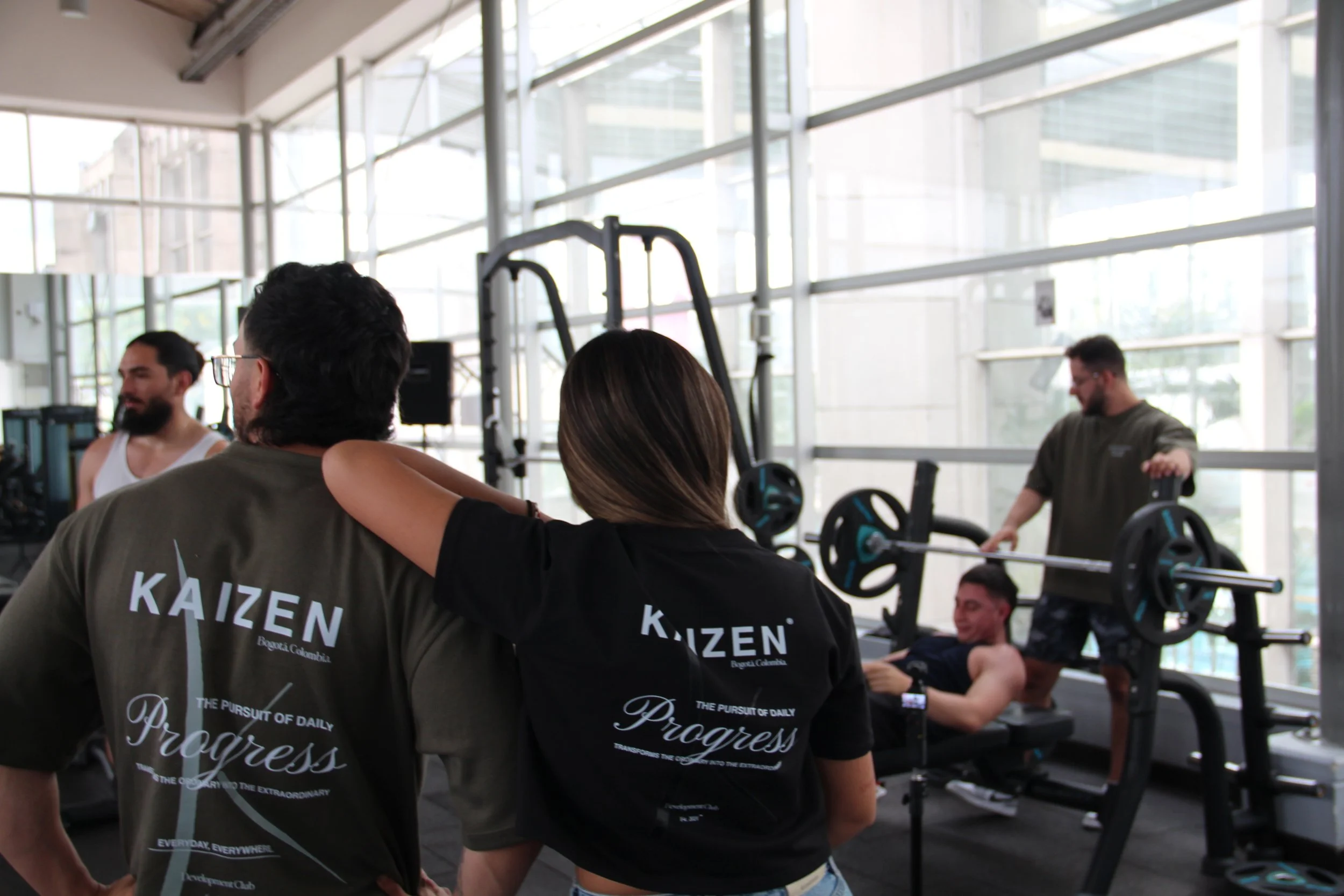 Personas en un gimnasio de entrenamiento de fuerza, dos mujeres en primer plano y dos hombres en el fondo, uno levantando pesas.