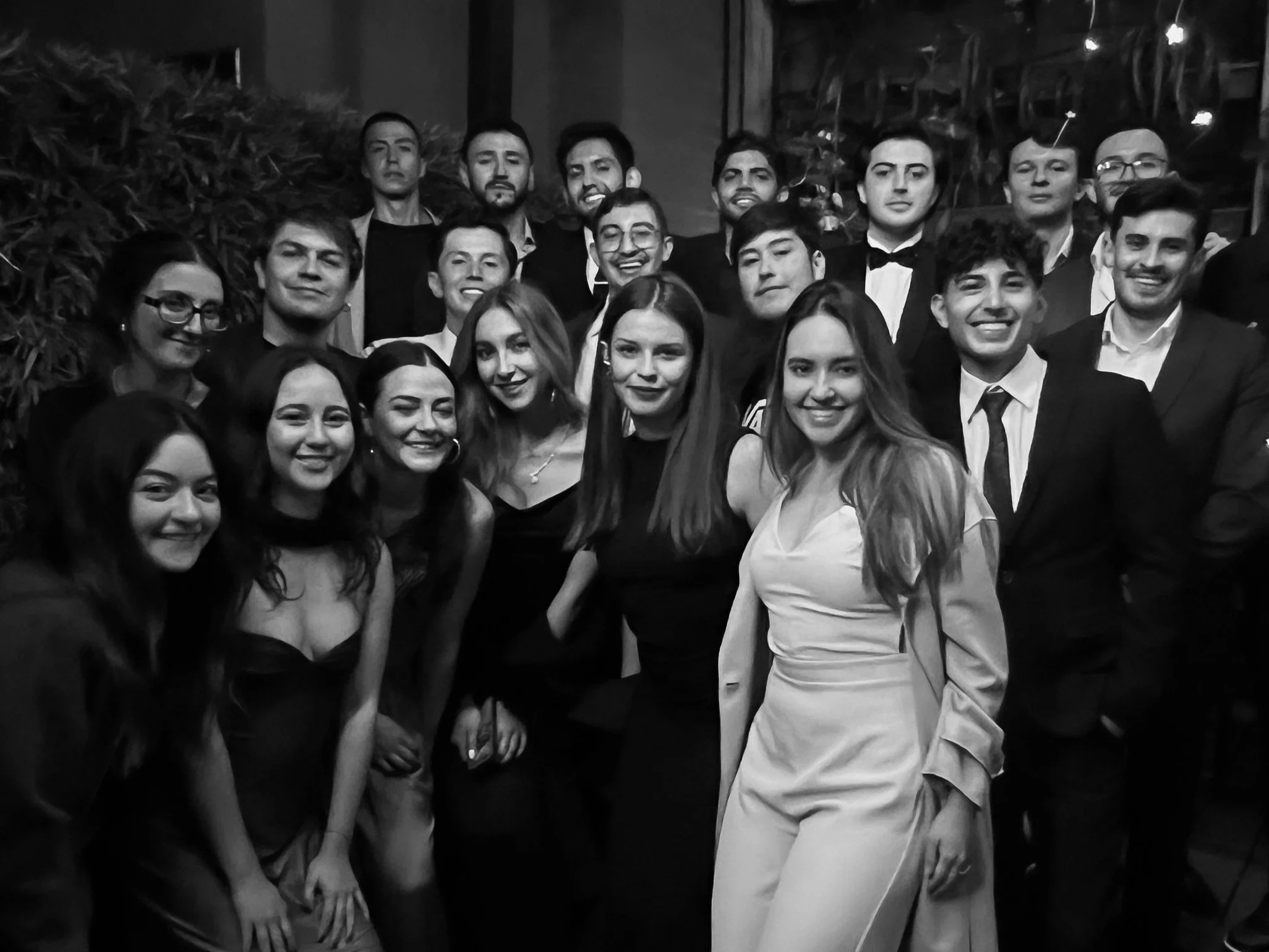 Grupo de personas en una celebración, muchas con ropa formal, en un lugar con decoración moderna y plantas.