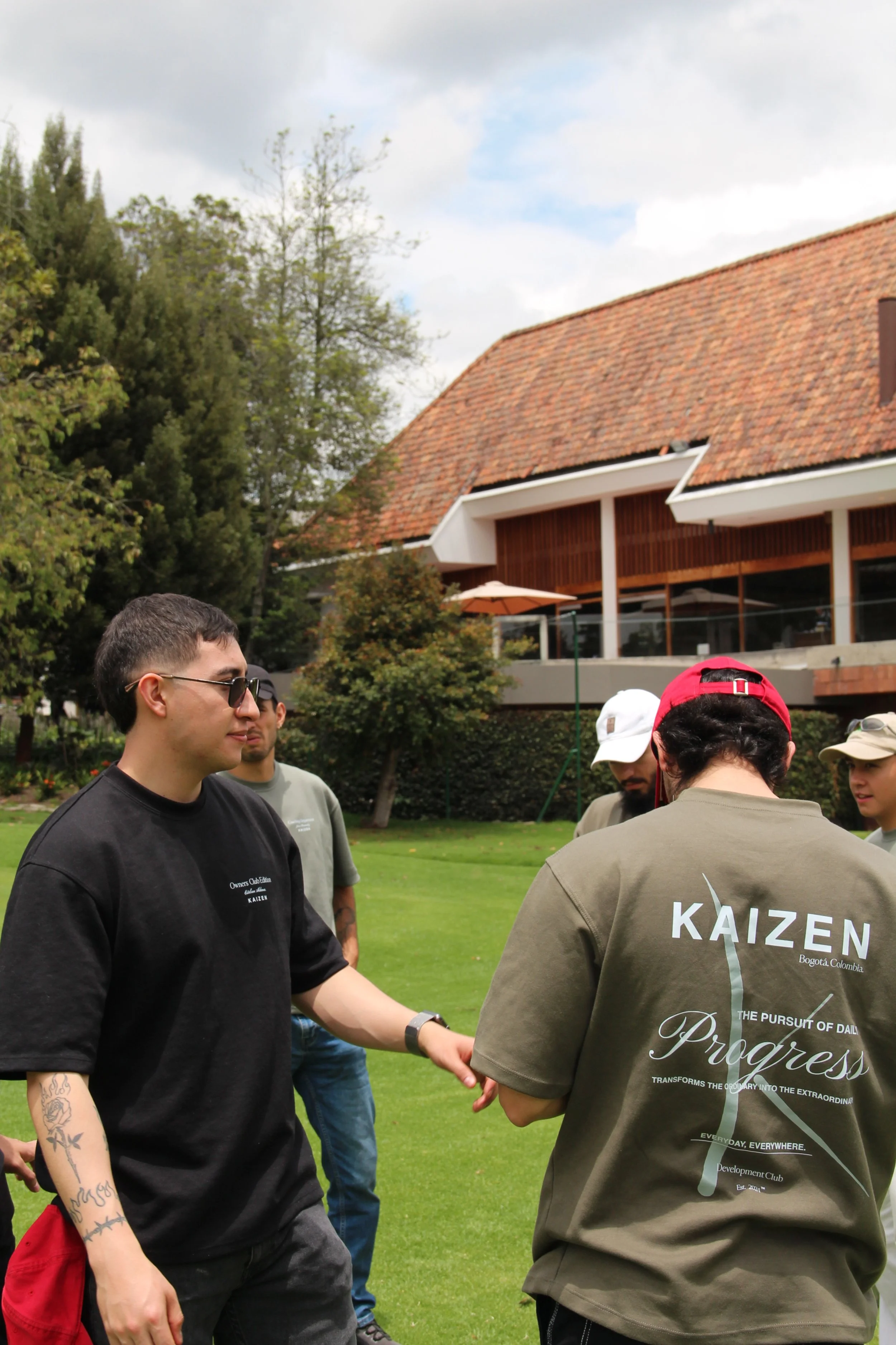 Grupo de jóvenes conversando en un jardín con césped verde y árboles, en un día soleado, en frente de una casa con techo de tejas rojas.