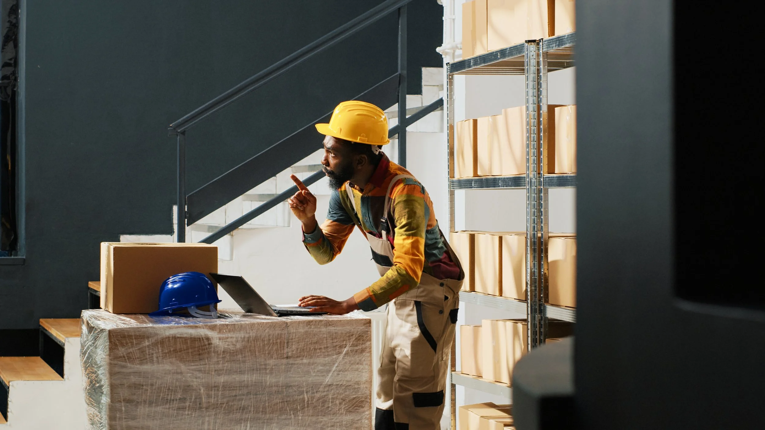 A construction worker wearing a yellow safety helmet, colorful shirt, and beige pants, standing at a desk with a laptop, pointing with their index finger, and appearing to be speaking or giving instructions. There are shelves with beige boxes behind him, and a blue safety helmet placed on the desk.