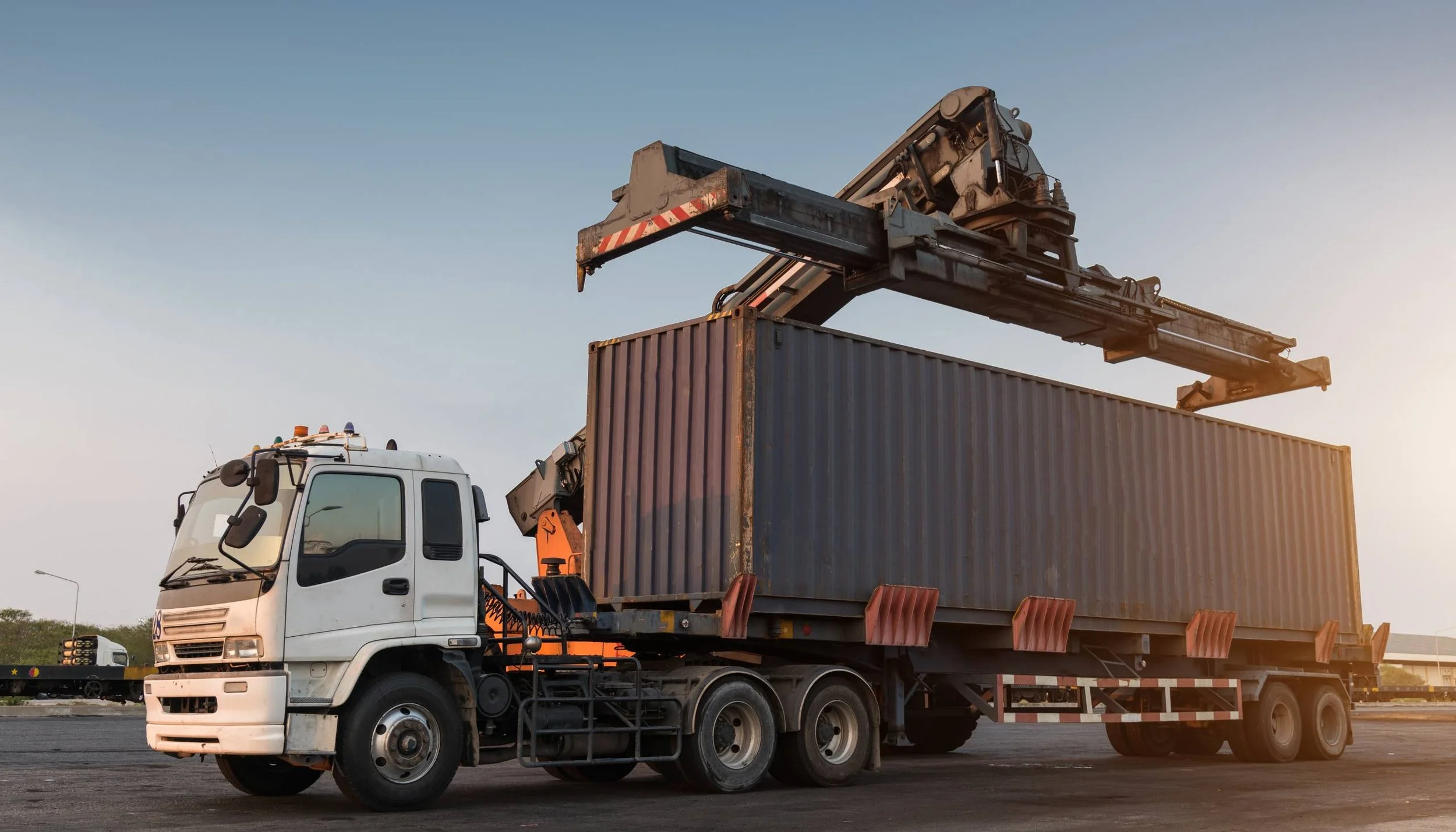 A cargo truck with a large shipping container on its trailer, being loaded with a crane at a port during sunset.