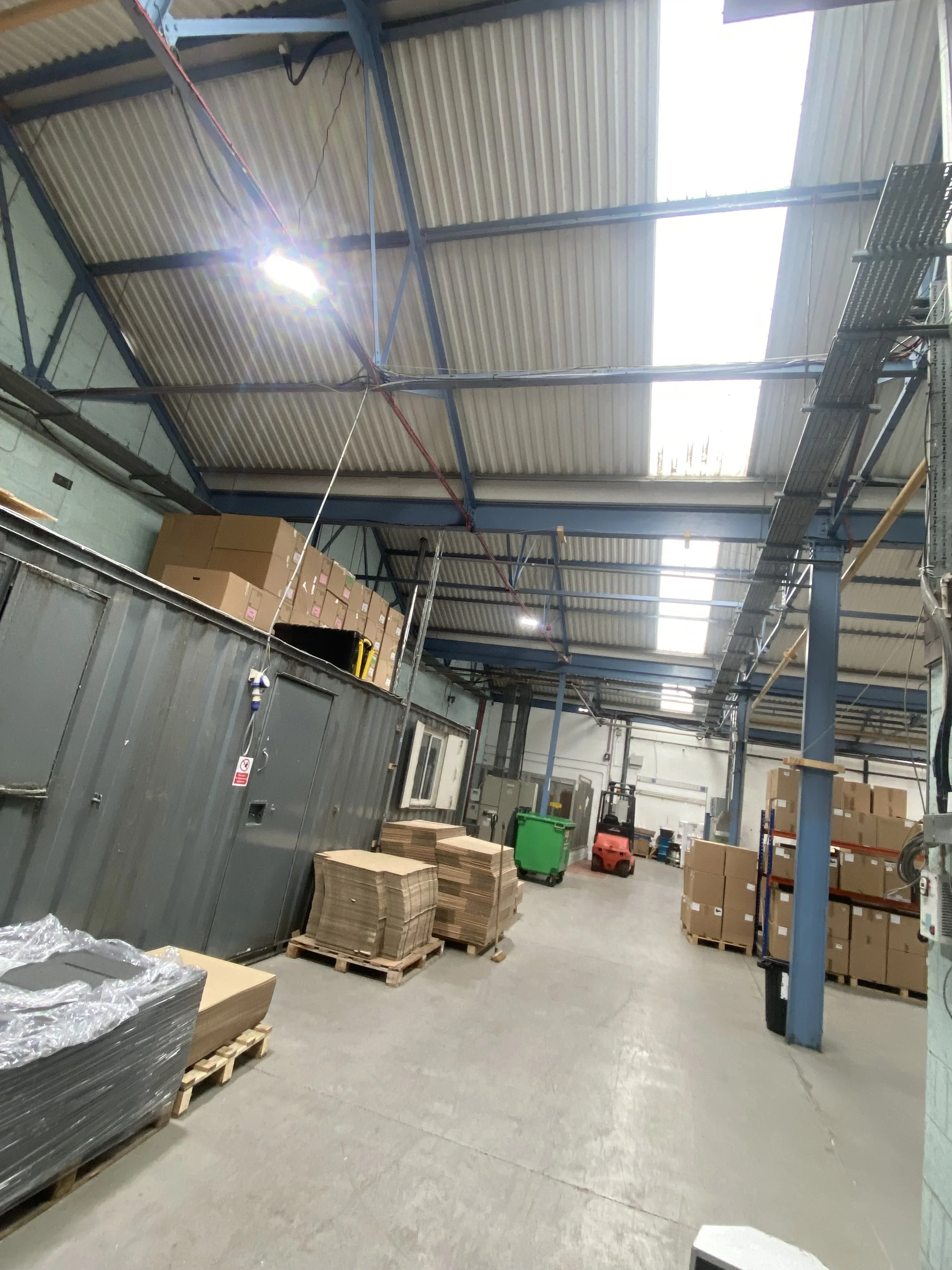 Warehouse aisle with pallets of flat-packed cardboard and stacked boxes, lit by overhead LED high-bay lights beneath a steel-framed roof with skylights
