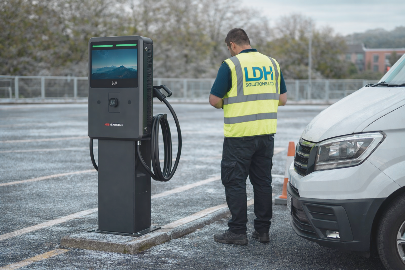 Outdoor DC EV charger installation in a commercial car park, with an electrician wearing a high-visibility vest completing works next to a pedestal-mounted charging unit on a frosty surface.