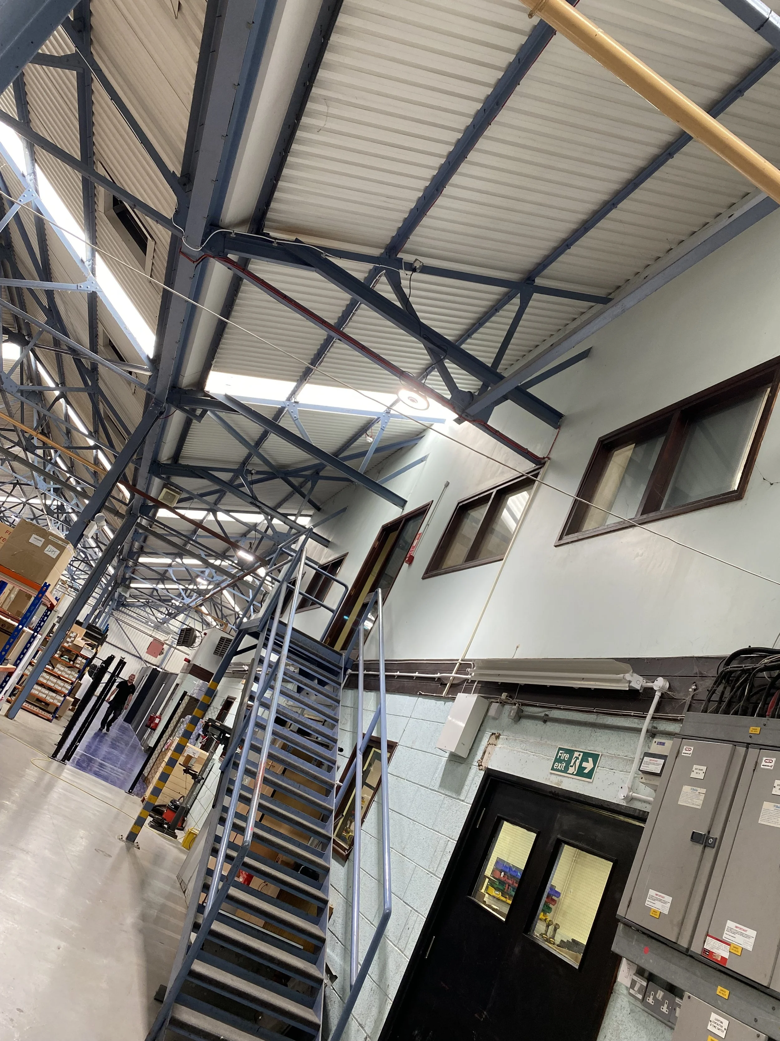 Long warehouse aisle beside mezzanine offices and a fire exit door, with suspended services and LED high-bay lights providing even illumination.