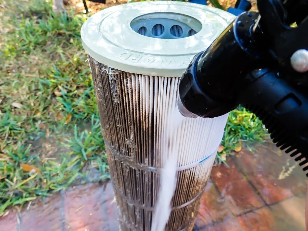 Person using a pressure washer to clean a cylindrical pool filter outdoors.