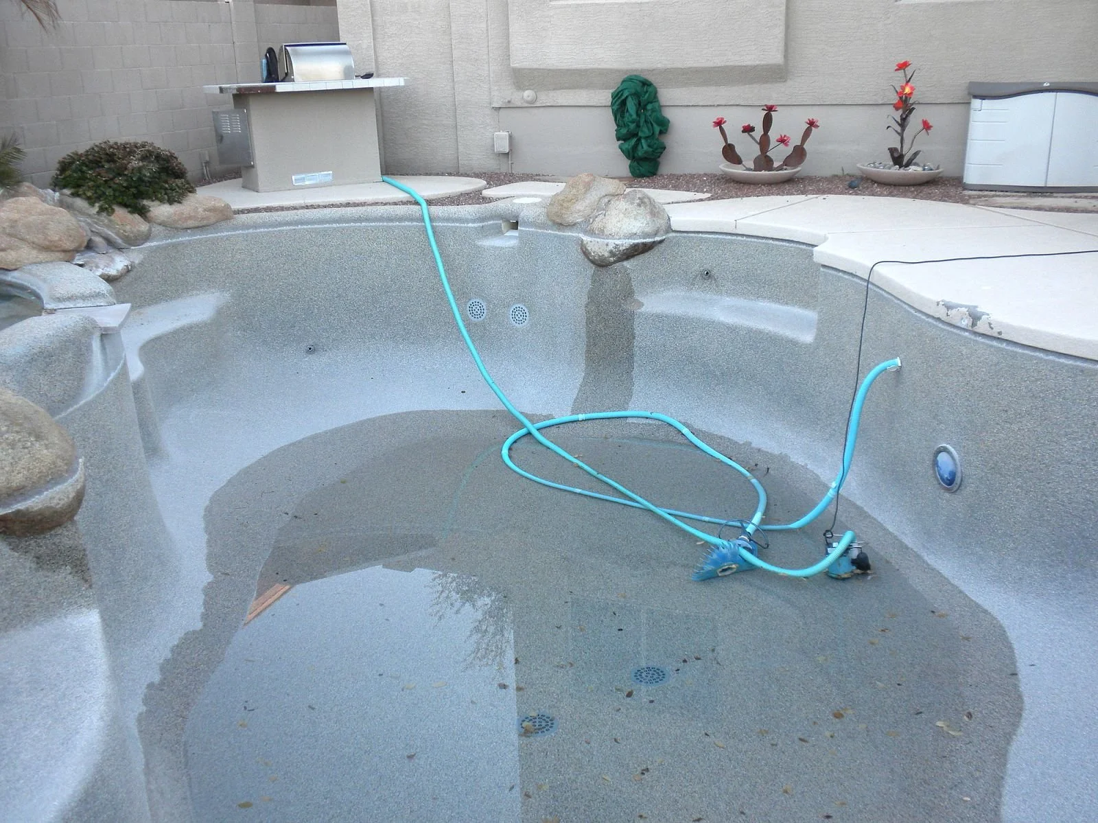 Empty swimming pool with blue pool cleaning equipment and hoses inside, surrounded by patio area with potted plants.