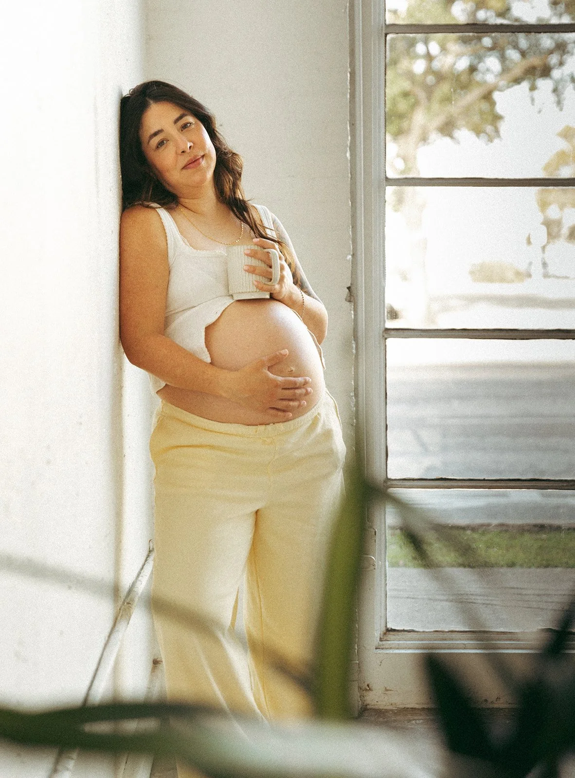A pregnant woman standing by a window, holding her baby bump with one hand and a mug with the other, dressed in a white sleeveless top and yellow pants, smiling softly.