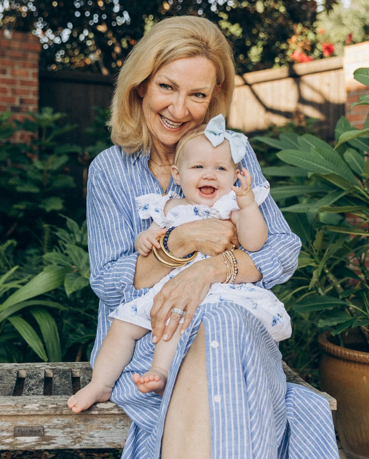 A smiling older woman with blonde hair holding a laughing baby girl with a big white bow in her hair in a garden with plants and a brick wall.