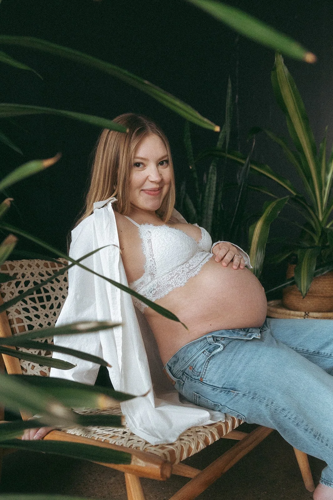 Pregnant woman sitting on a chair surrounded by green plants, wearing a white lace top and light blue jeans, smiling at the camera.