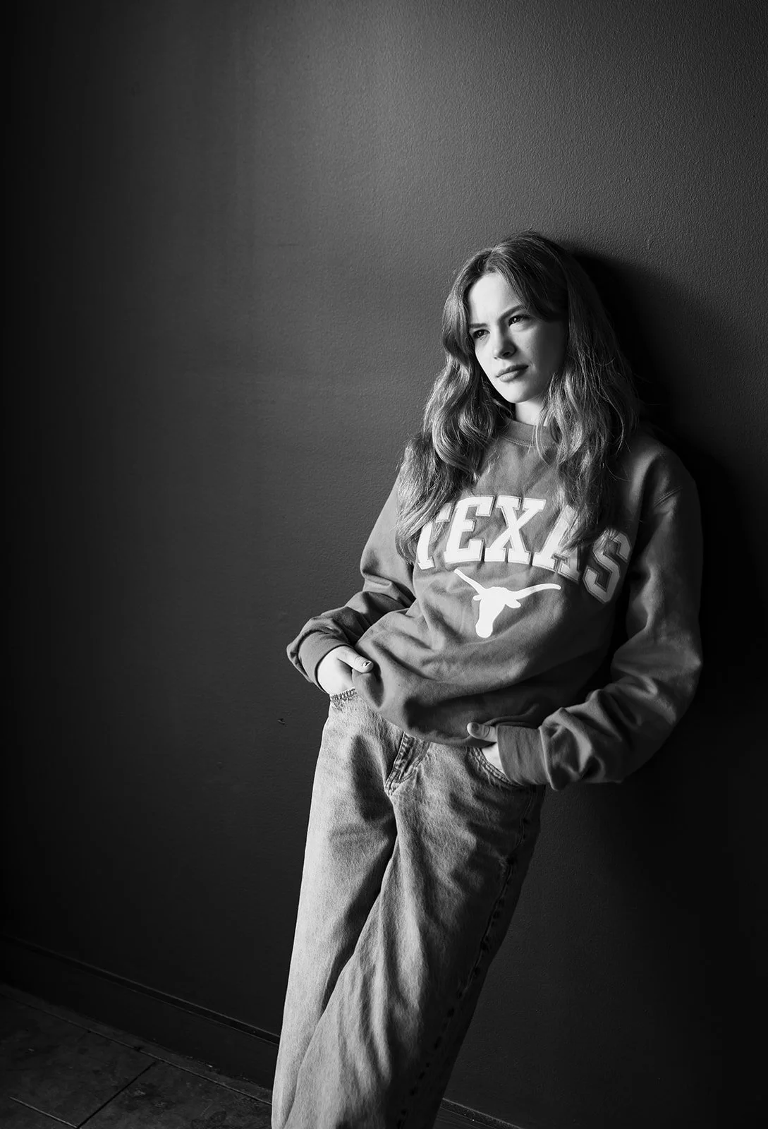 A young woman with wavy hair leaning against a wall in a casual outfit, wearing a sweatshirt that says 'TEXAS' and jeans.