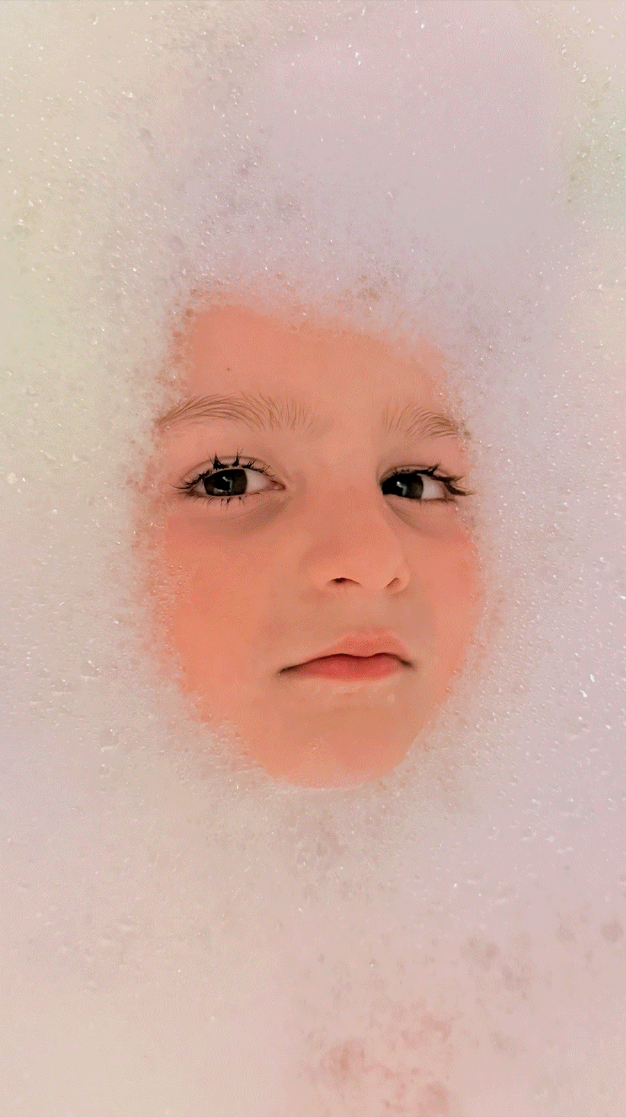 Close-up of a child's face with fair skin and dark eyes, submerged in a bath with white bubbles surrounding the face.