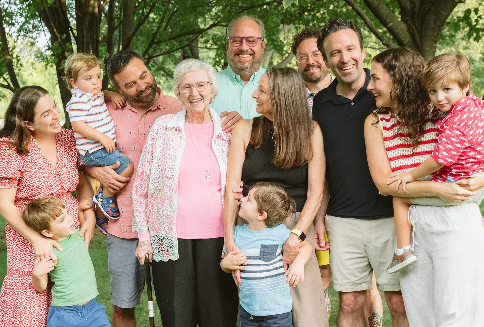 A multigenerational family of adults and children gathering outdoors in a park for a group photo, smiling and interacting.