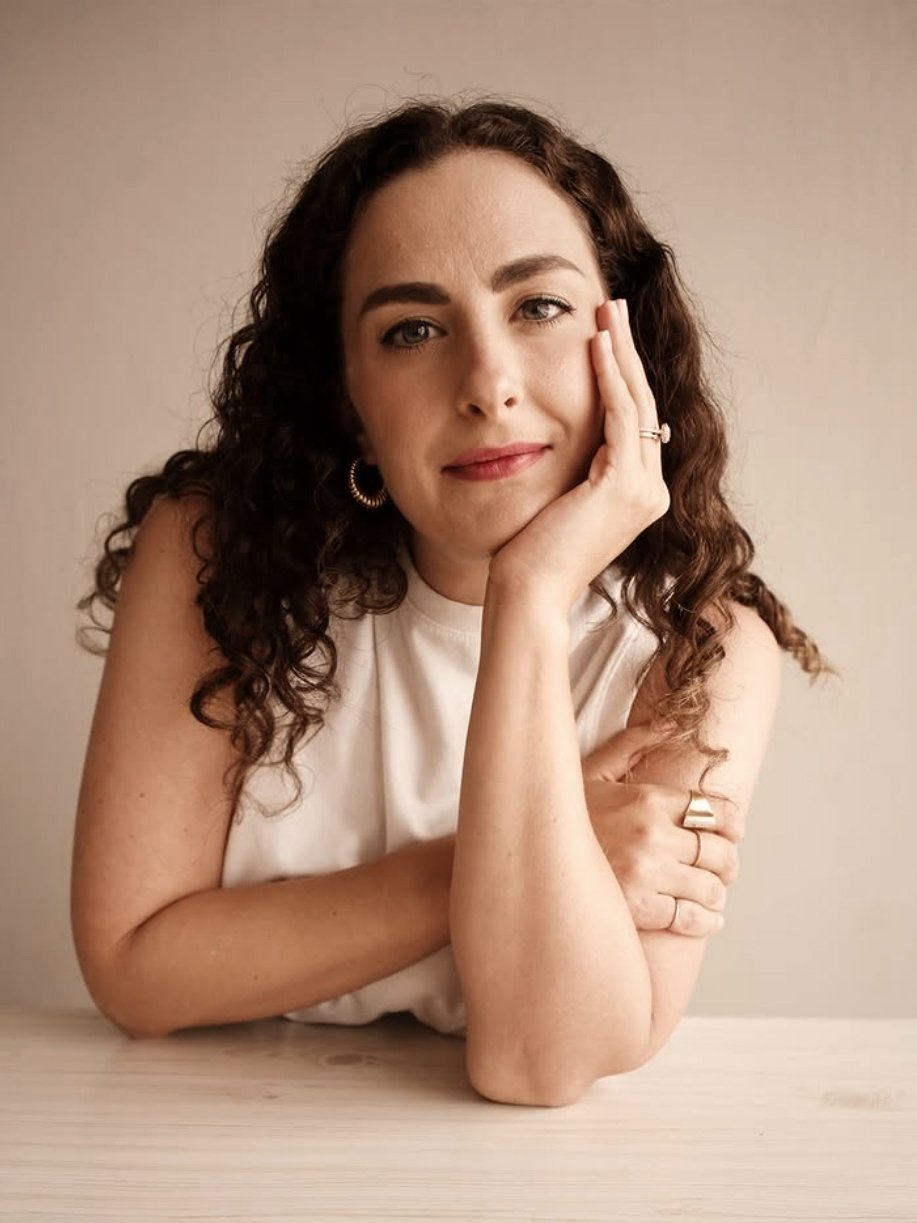 A woman with curly brown hair resting her head on her left hand, sitting at a light-colored table, wearing a sleeveless white top, gold hoop earrings, and rings, with a neutral beige background.