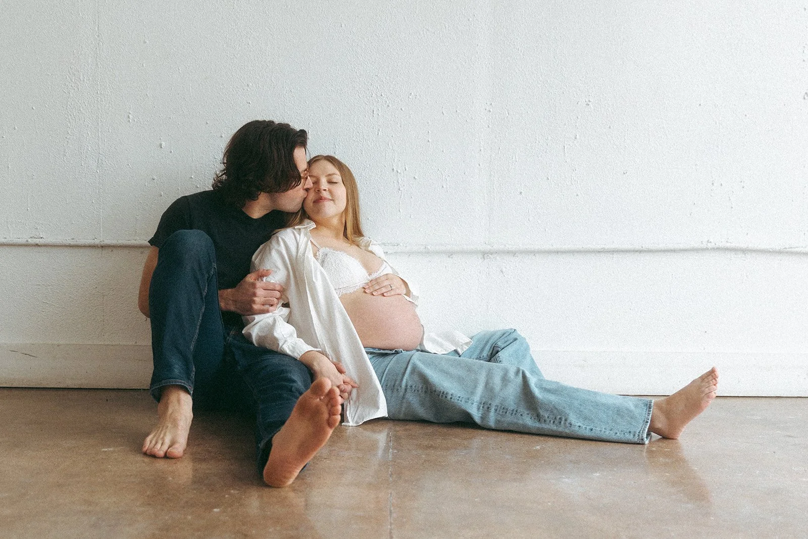 A pregnant woman sitting on the floor with her legs extended, wearing a white shirt and jeans, with a man beside her, both interacting affectionately against a plain white wall.