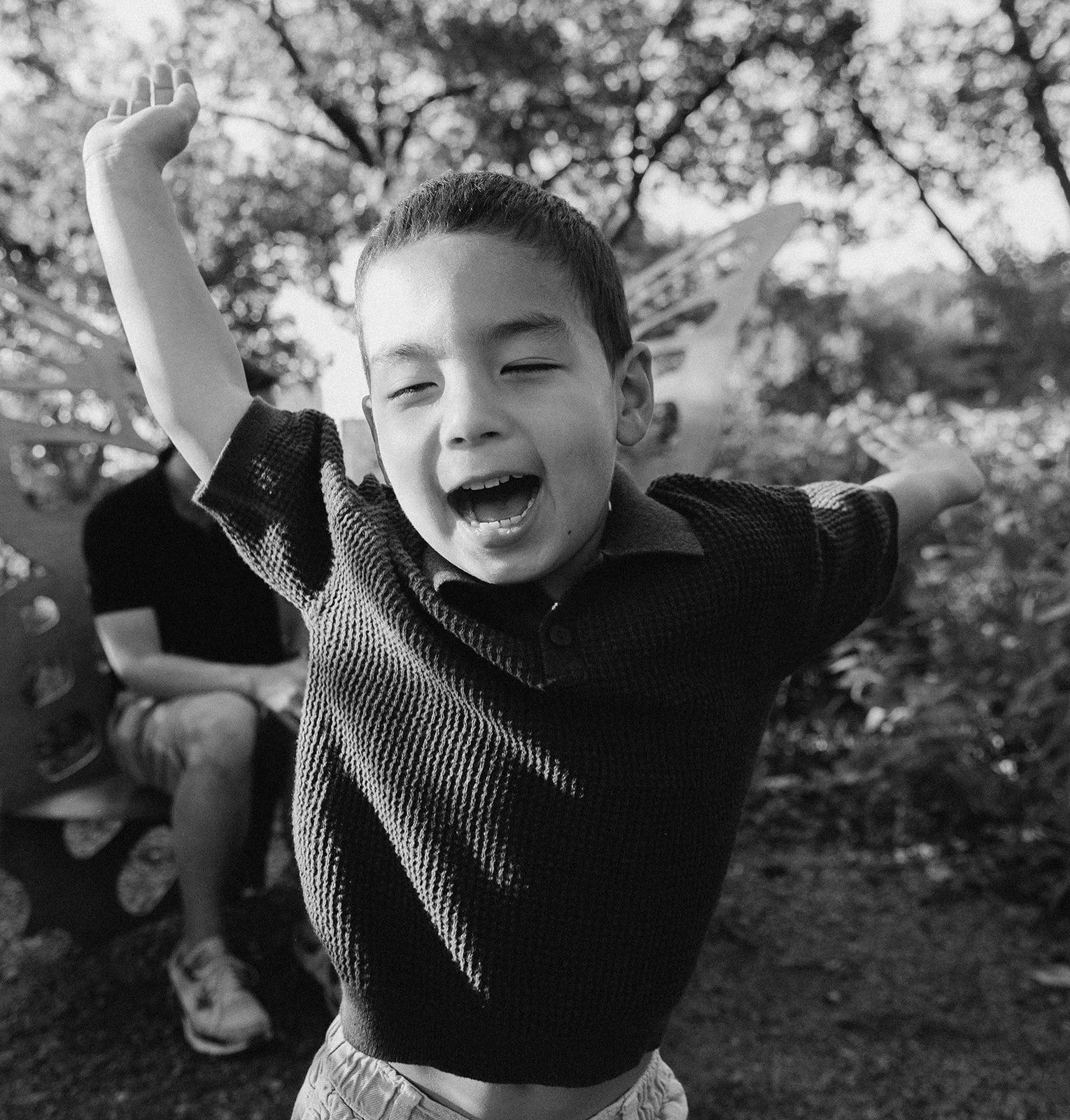 A young boy with short hair, smiling with eyes closed, raising his arms, outdoors with trees and bushes in the background, in black and white.