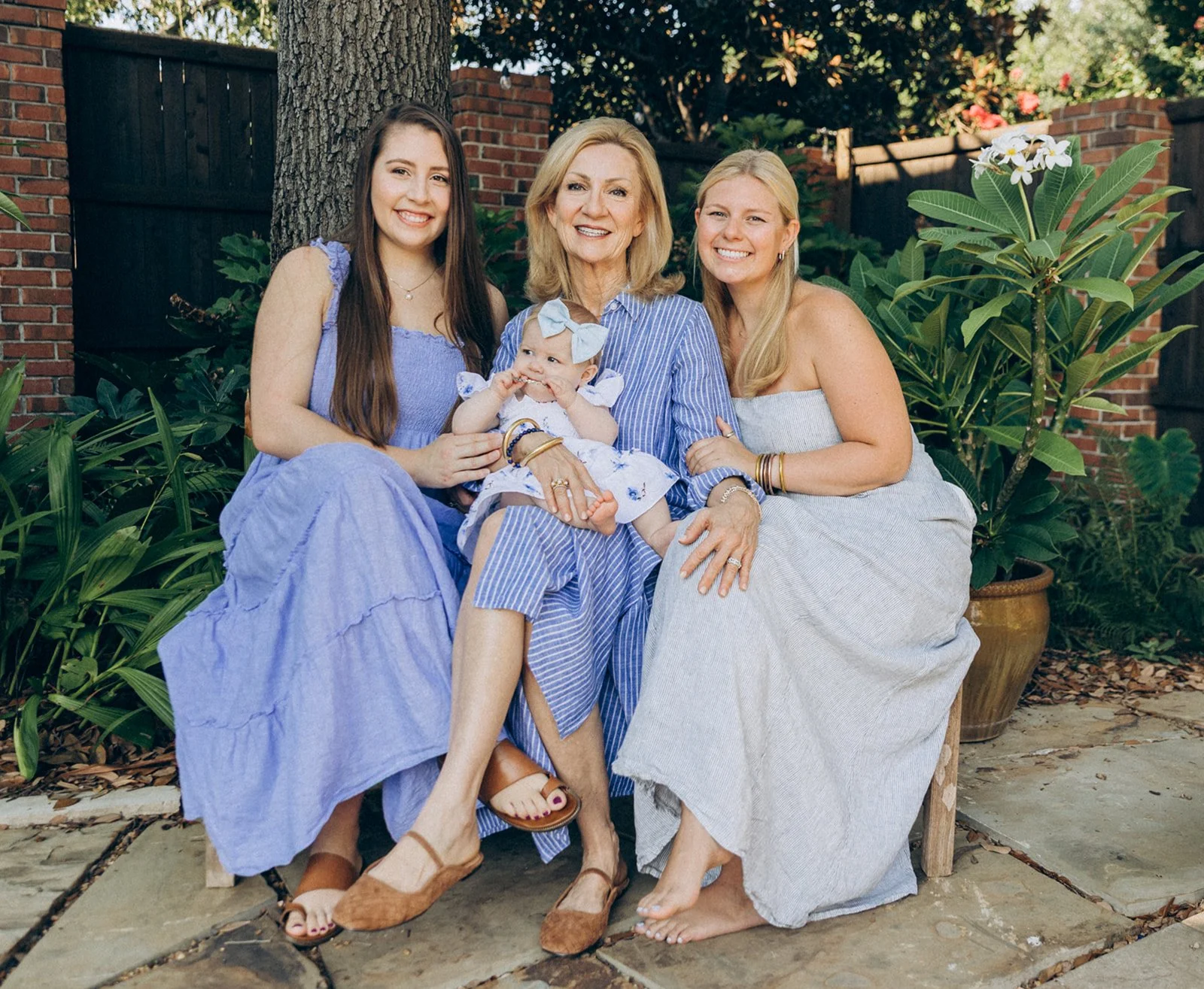Four women, including an elderly woman and three younger women, sitting outdoors on a bench with a baby, surrounded by green plants, brick wall, and trees.