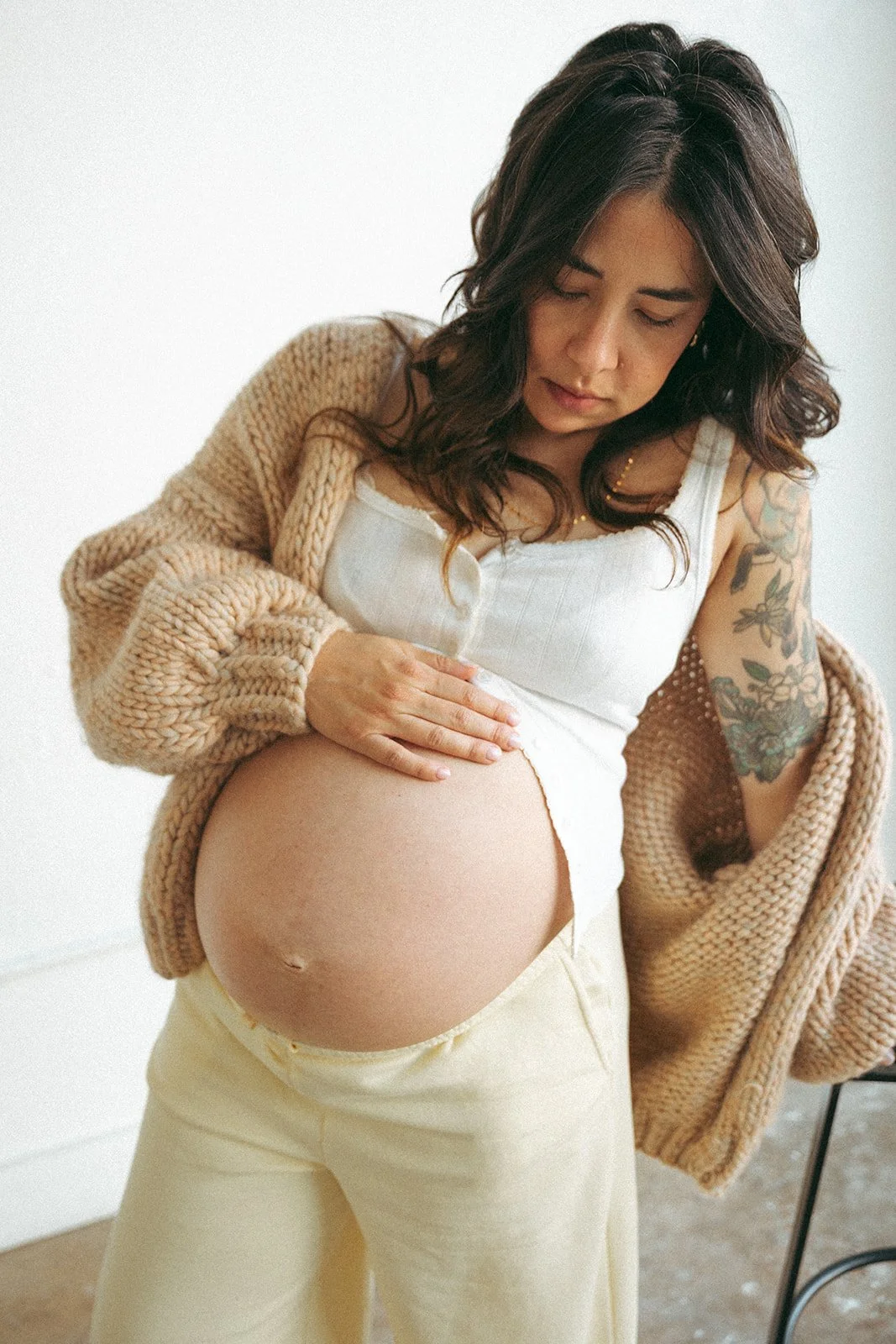 A woman with dark, wavy hair and tattoos on her arm looks down at her pregnant belly while holding it with one hand. She is wearing a white tank top, light-colored pants, and a chunky beige cardigan, standing against a plain white wall.