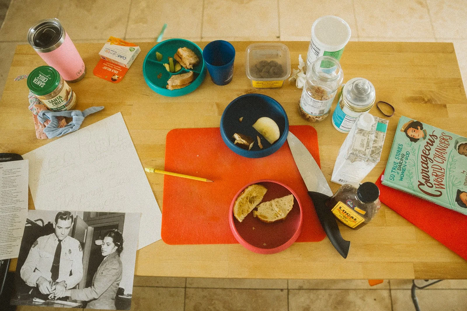 A cluttered wooden table with various items including food containers, pills, a knife, a book titled 'Nourishing Wounded Changemakers,' a black-and-white photograph of a man and woman, a notebook with handwritten notes, and a few bottles and cups.
