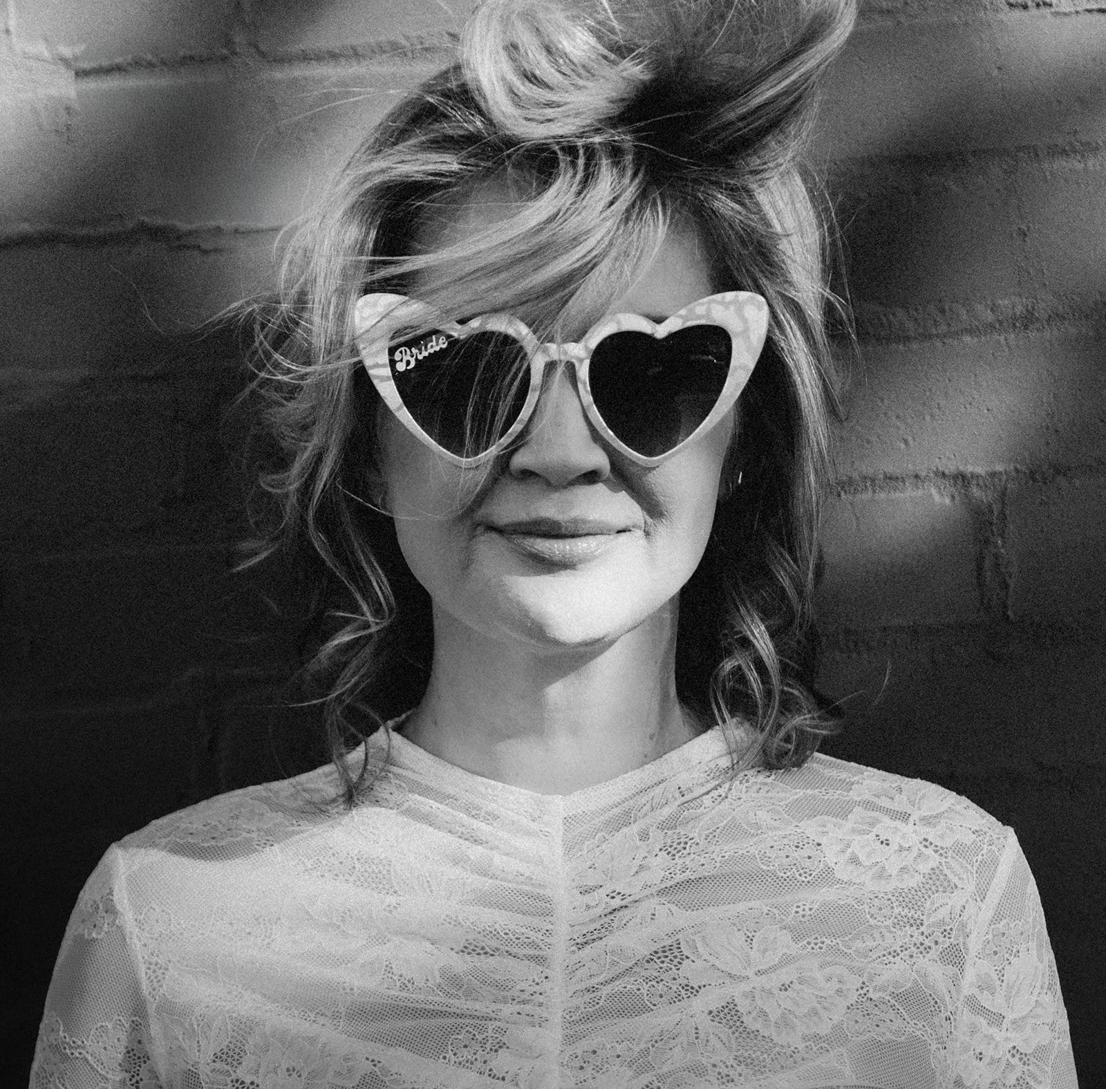 A woman with curly hair wearing heart-shaped sunglasses and a lace top, smiling against a brick wall.