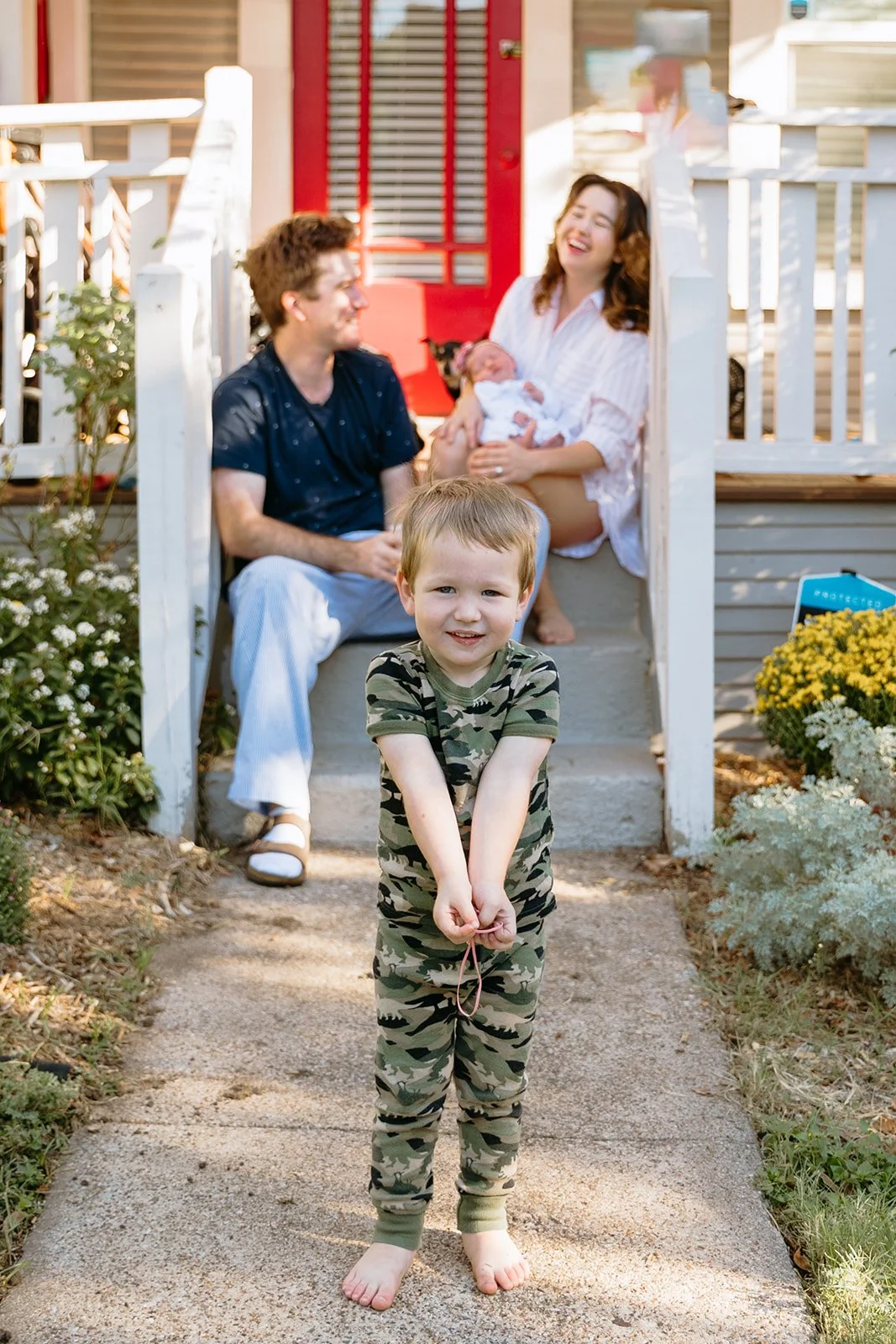A young boy in camouflage pajamas stands on the sidewalk in front of a house, smiling at the camera. Behind him on the porch, a family of four is sitting and enjoying time together, with the mother holding a newborn, the father smiling, and another c