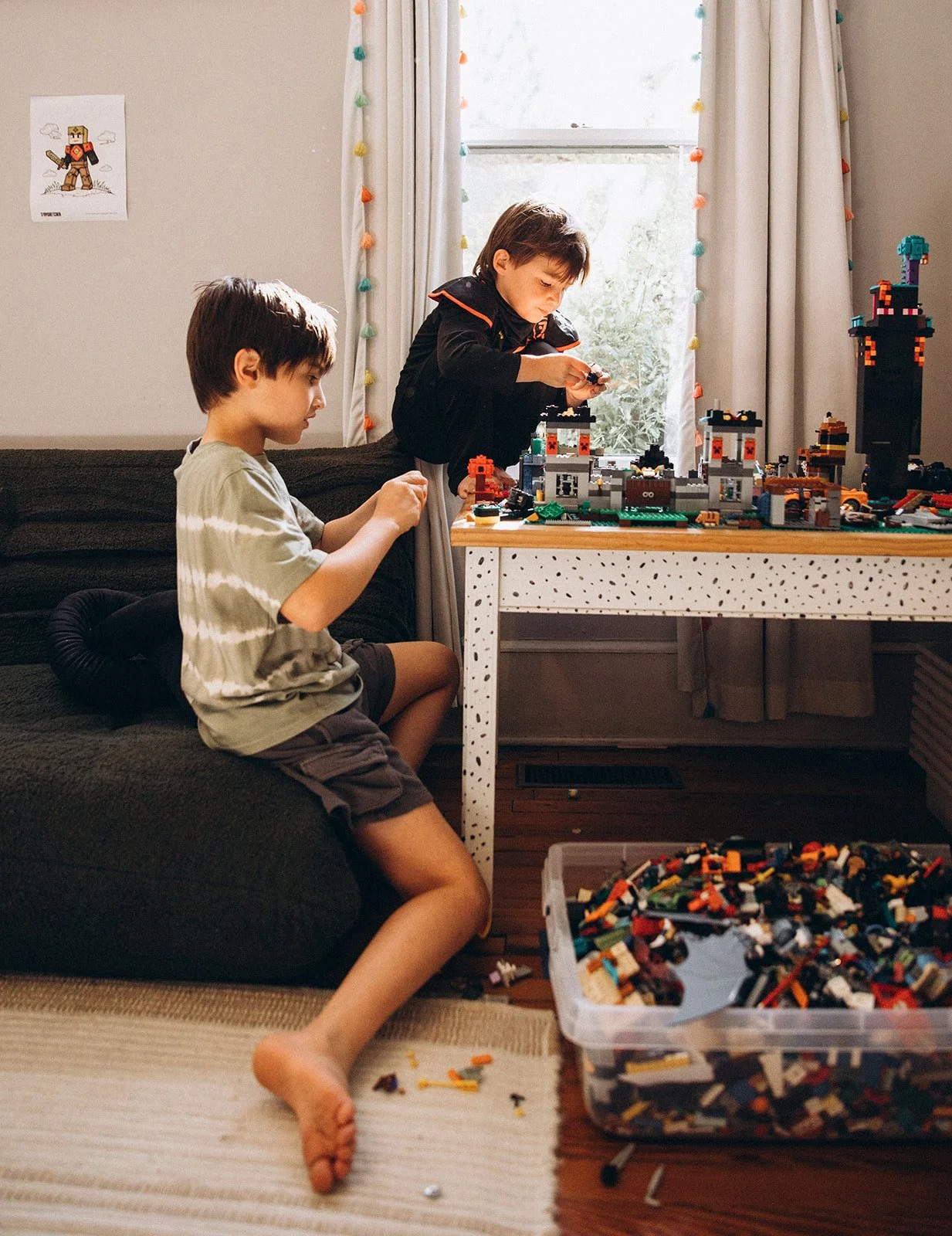 Two young boys playing with LEGO bricks in a cozy room, one sitting on a couch and the other standing on a table, with a large container of LEGO pieces nearby.