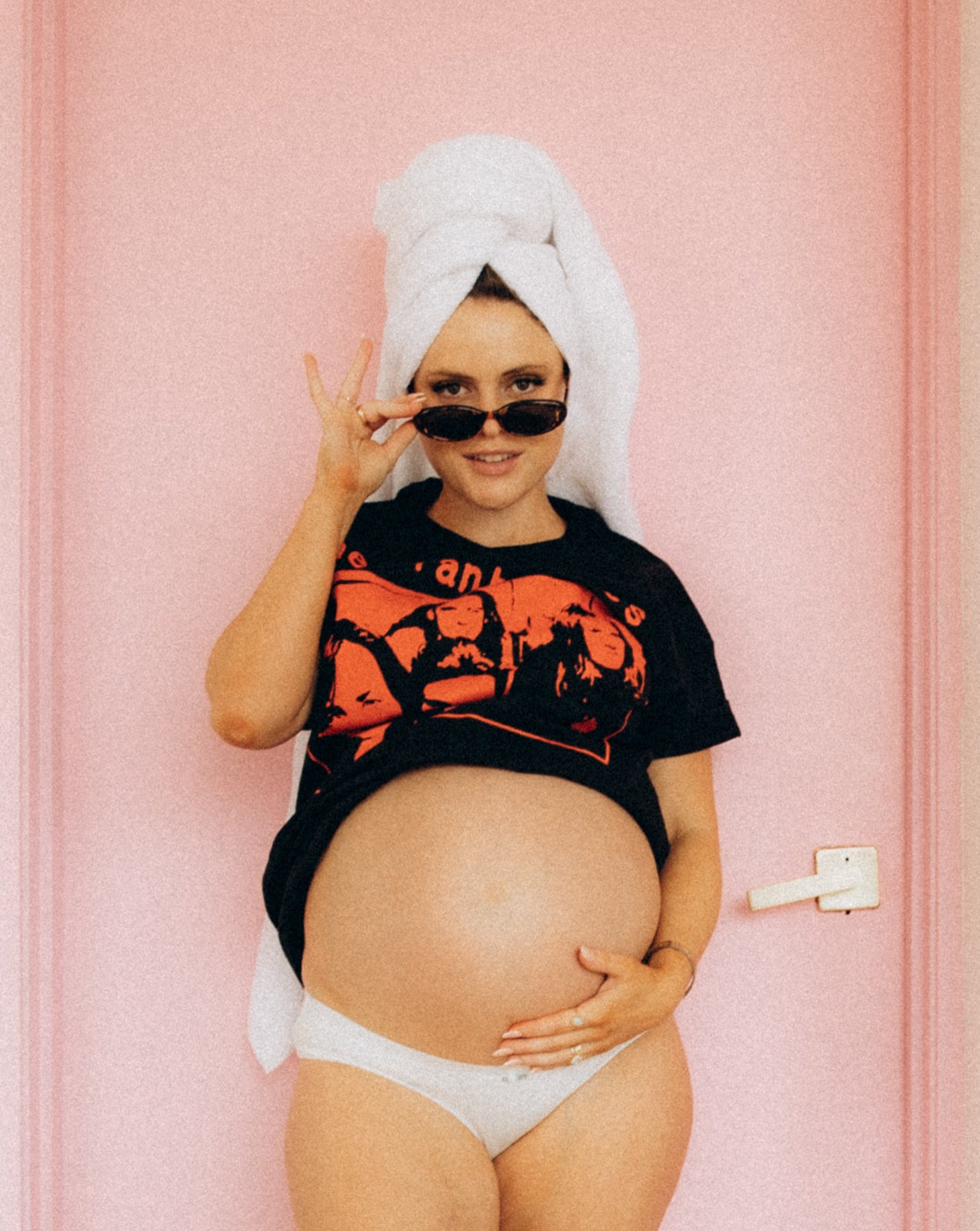 Pregnant woman with towel wrapped around her head, wearing sunglasses, black graphic t-shirt, and white underwear, standing against pink wall.