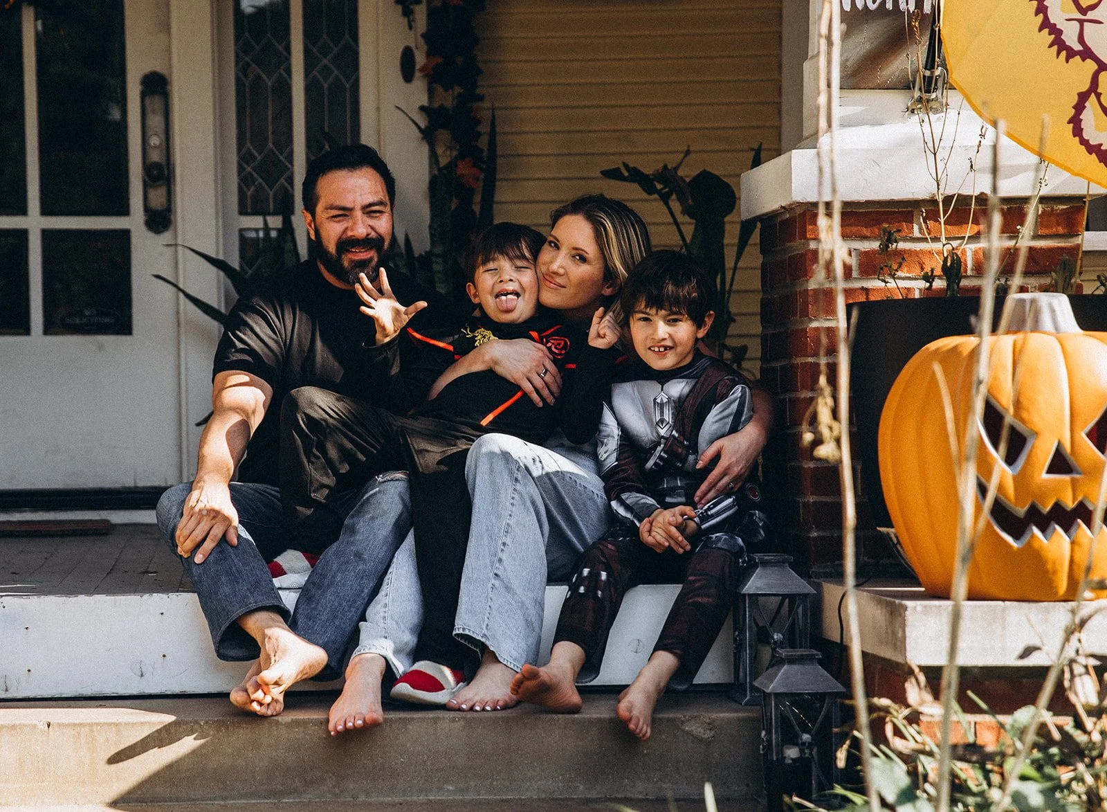 A family of four sitting on their front porch dressed in Halloween costumes, with jack-o'-lantern decorations nearby, celebrating Halloween.