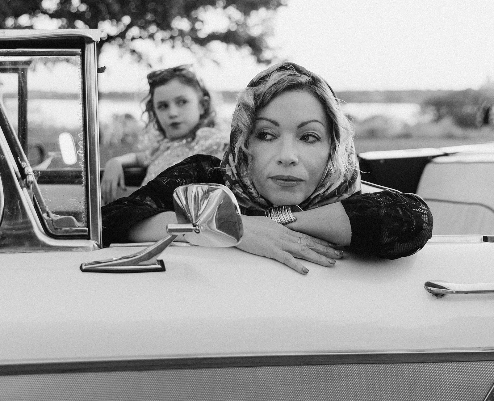 A woman with wavy blonde hair leaning on the hood of a vintage convertible car, resting her chin on her crossed arms while looking away. A young girl with curly hair is sitting in the back seat, looking forward. The background features open fields an
