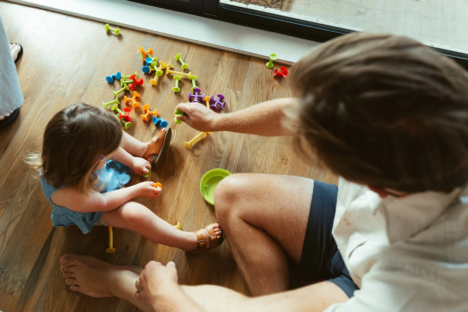 An adult and a young girl sitting on a wooden floor near a glass door, playing with colorful Tegu magnetic building blocks, with some on the floor and others held by the girl and the adult.