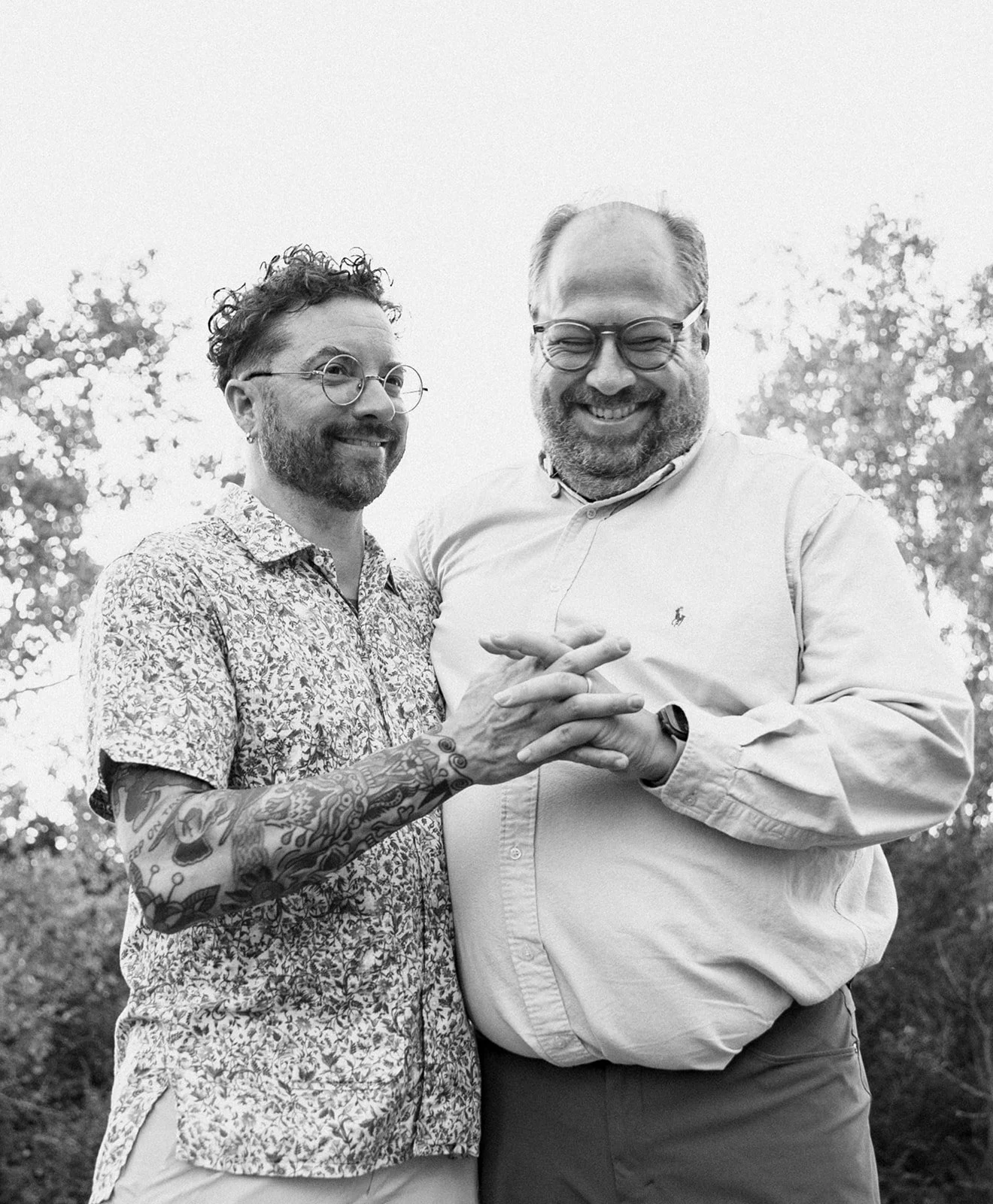 Two smiling men with glasses, one with tattoos, standing outdoors among trees, holding hands and looking happy.