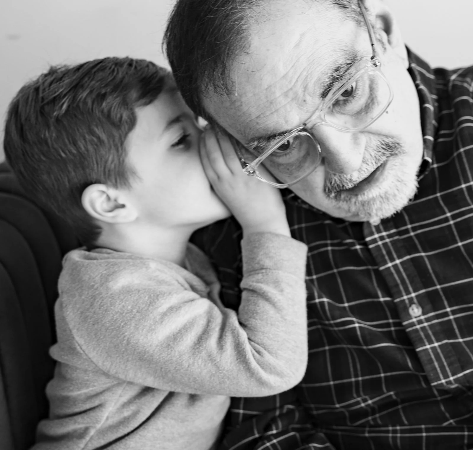 A young boy whispering into an older man's ear, who is wearing glasses, both with focused expressions. The photo is in black and white.