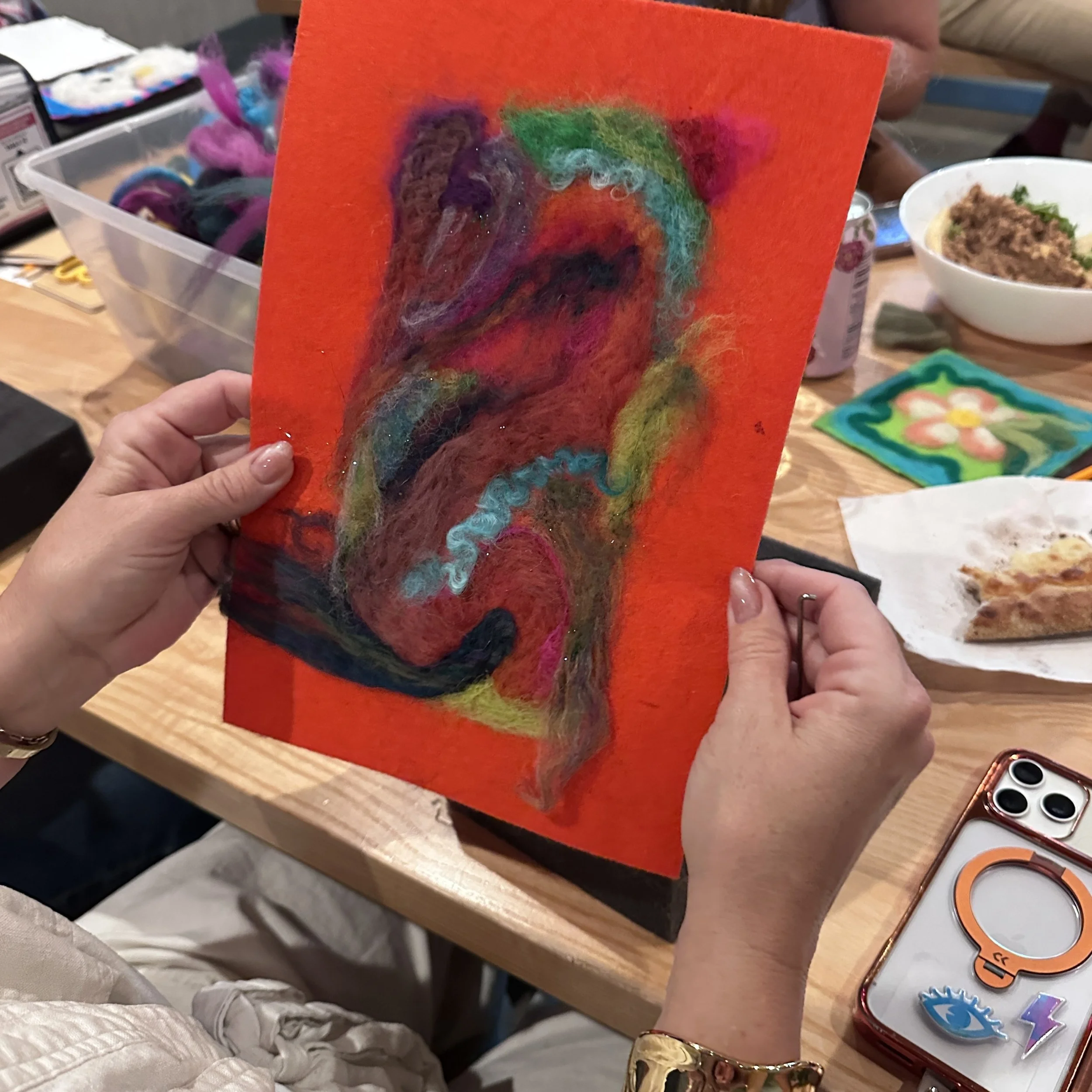 A person is holding a colorful felted artwork depicting a face with rainbow hair and dark features on an orange background. The setting includes craft supplies, yarn, and a bowl of food on a table.