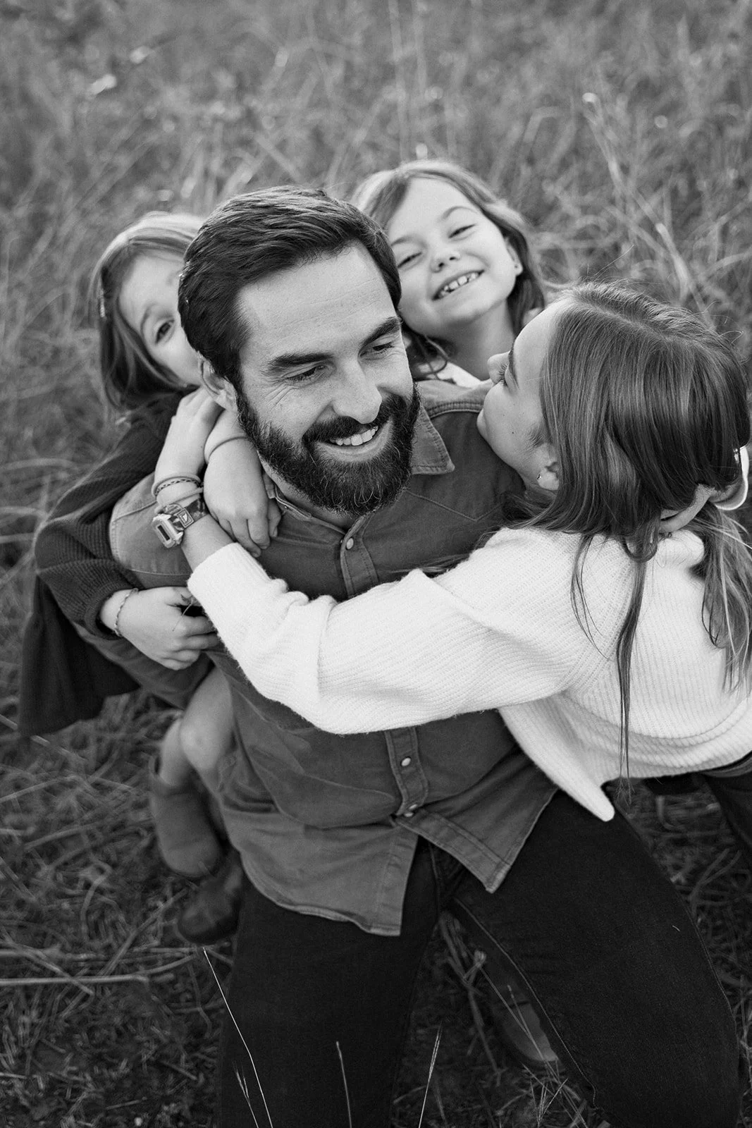 A man with a beard and three young girls outdoors, sharing a joyful embrace.